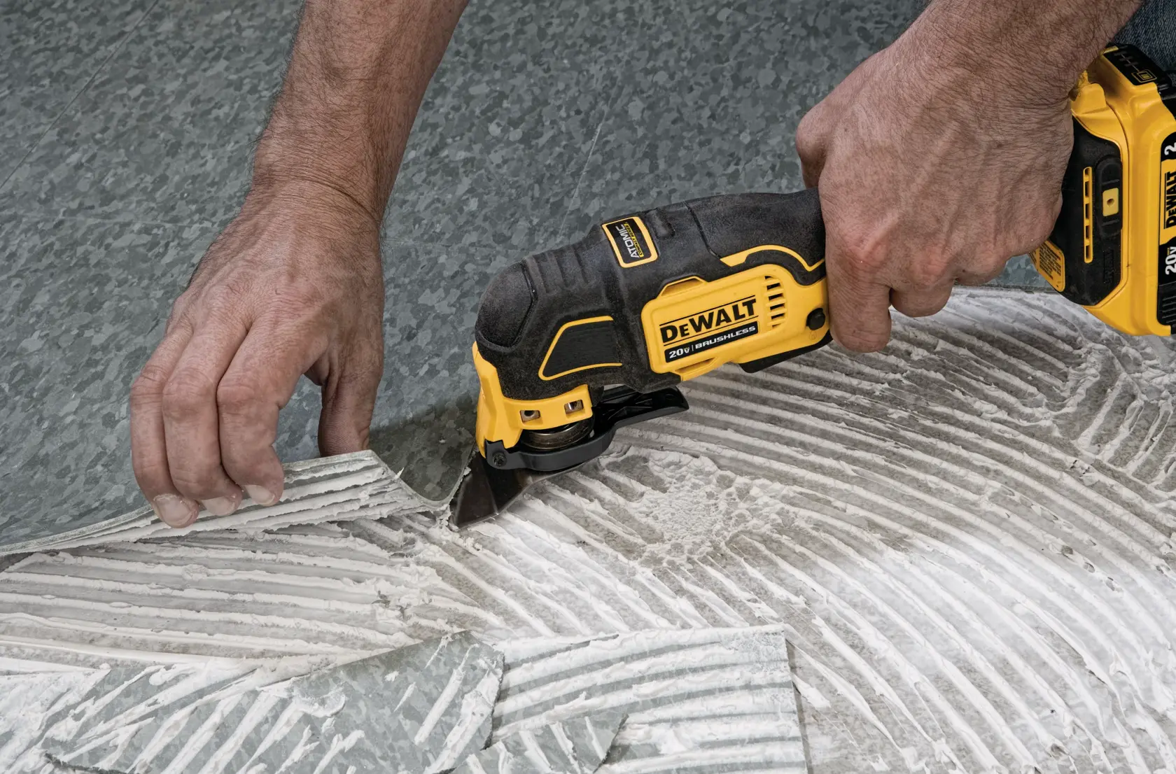 A DEWALT cordless oscillating multi-tool is being used to cut and remove flooring material. A person's hands are guiding the tool and lifting the material.