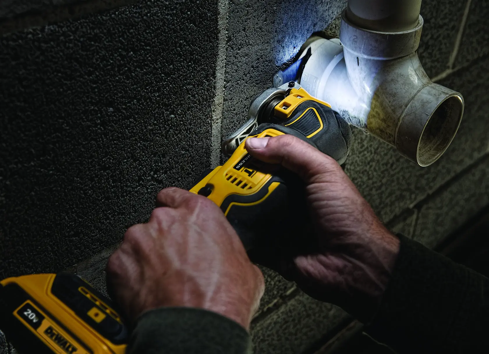 Close-up of hands using a DEWALT cordless oscillating multi-tool to cut into a concrete wall near a plastic plumbing pipe.