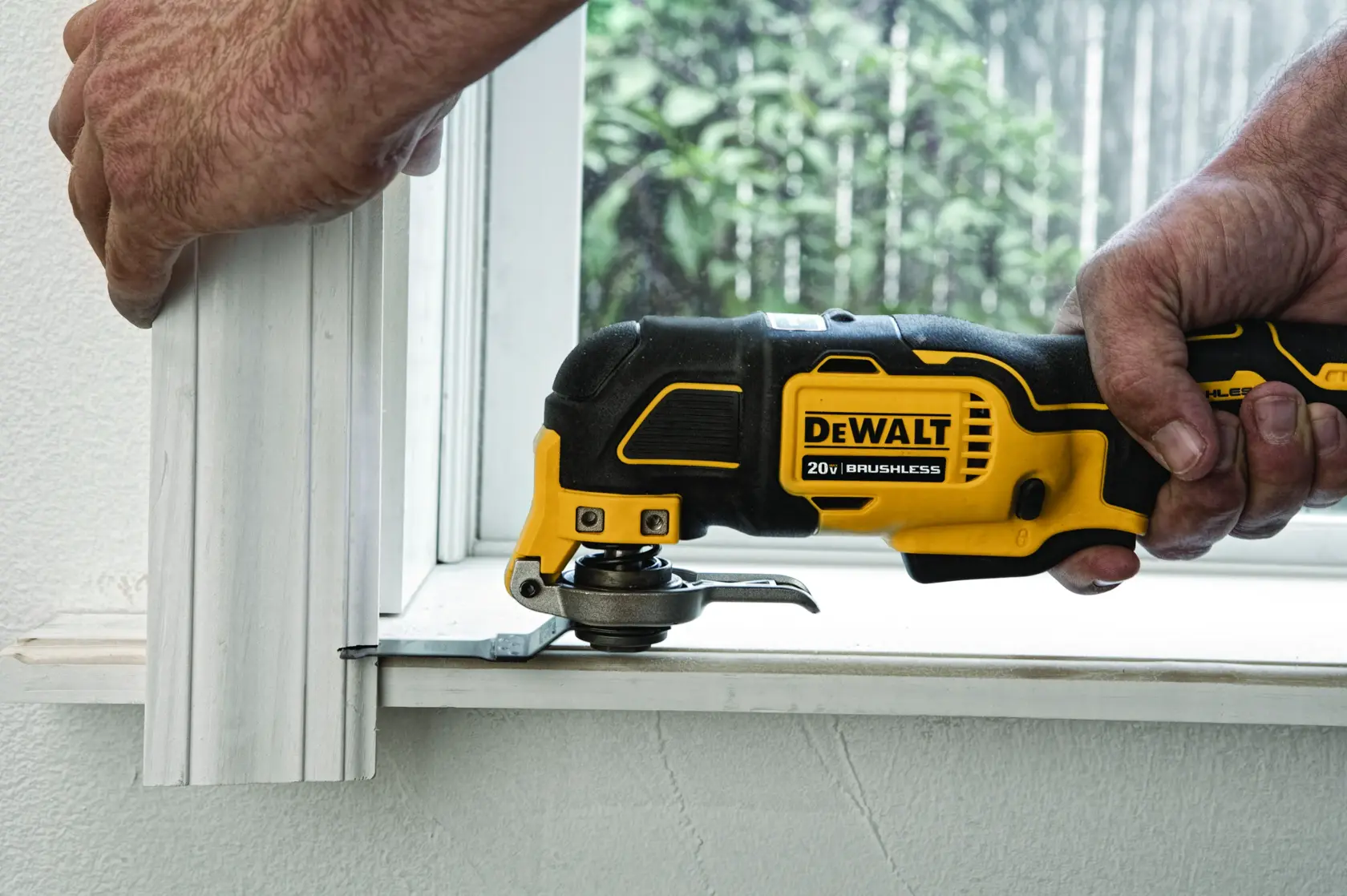 A DEWALT 20V brushless oscillating multi-tool being used to cut the bottom of a wooden trim near a window.