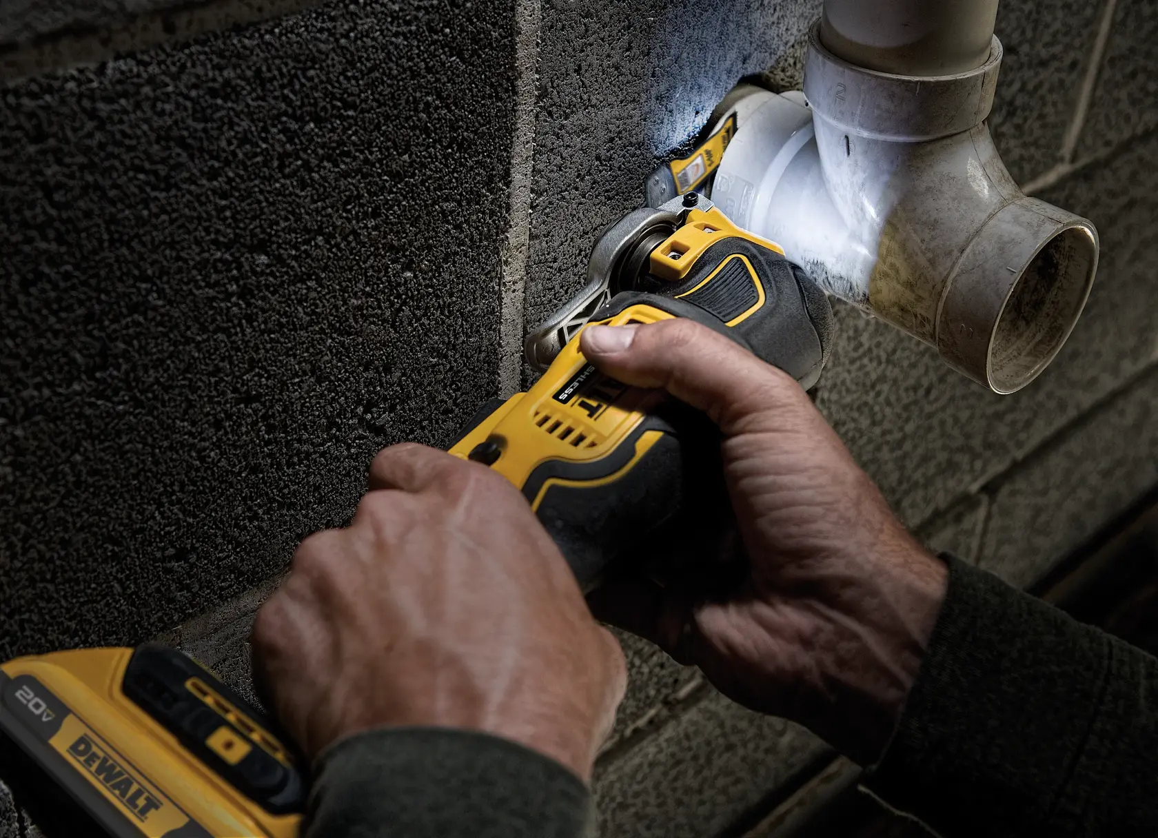 A person is using a yellow and black DEWALT 20V cordless oscillating multi-tool to cut or shape a piece of white plastic pipe attached to a concrete block wall.