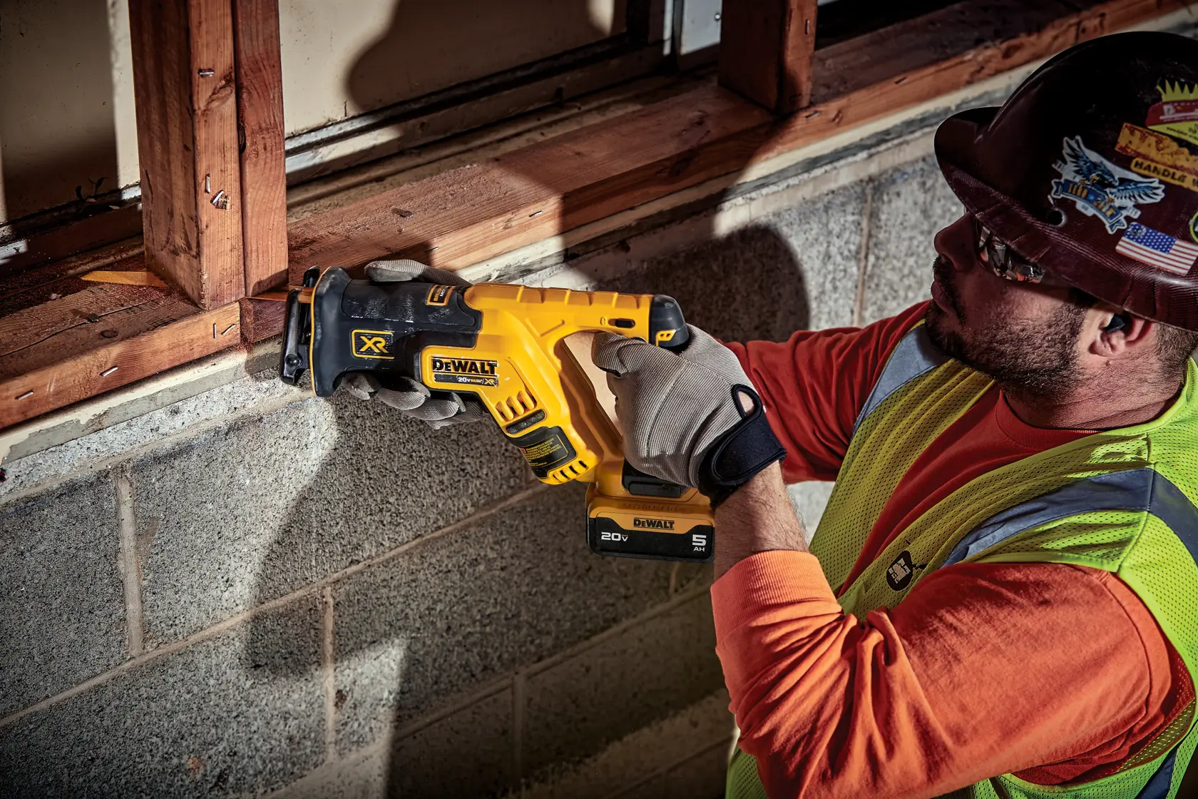 A construction worker wearing a safety vest and gloves uses a DEWALT cordless reciprocating saw to cut a wooden beam near a concrete wall. The worker's face is blurred for privacy.