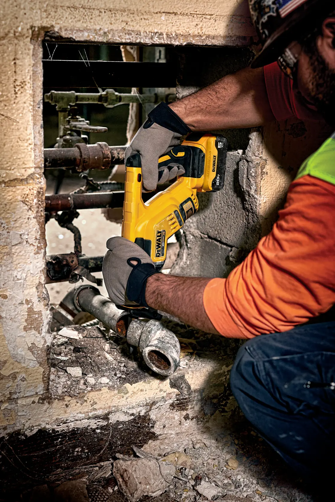 A person wearing work gloves and construction clothing uses a DEWALT cordless reciprocating saw to cut a metal pipe in a rough, partially demolished wall.