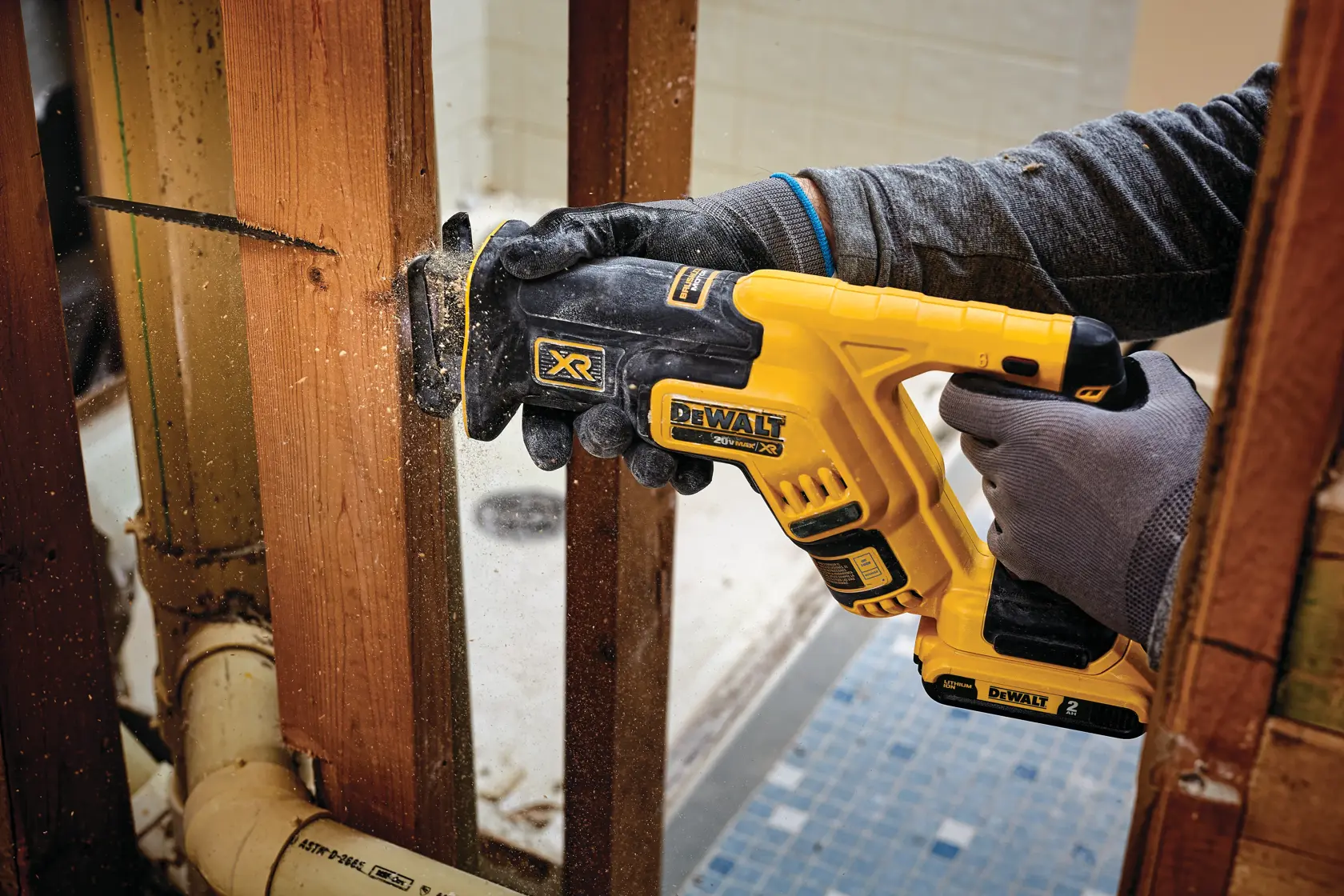 A person wearing gloves is using a yellow and black DEWALT cordless reciprocating saw to cut through a wooden beam during construction or renovation. Sawdust is visible as the tool cuts the wood.