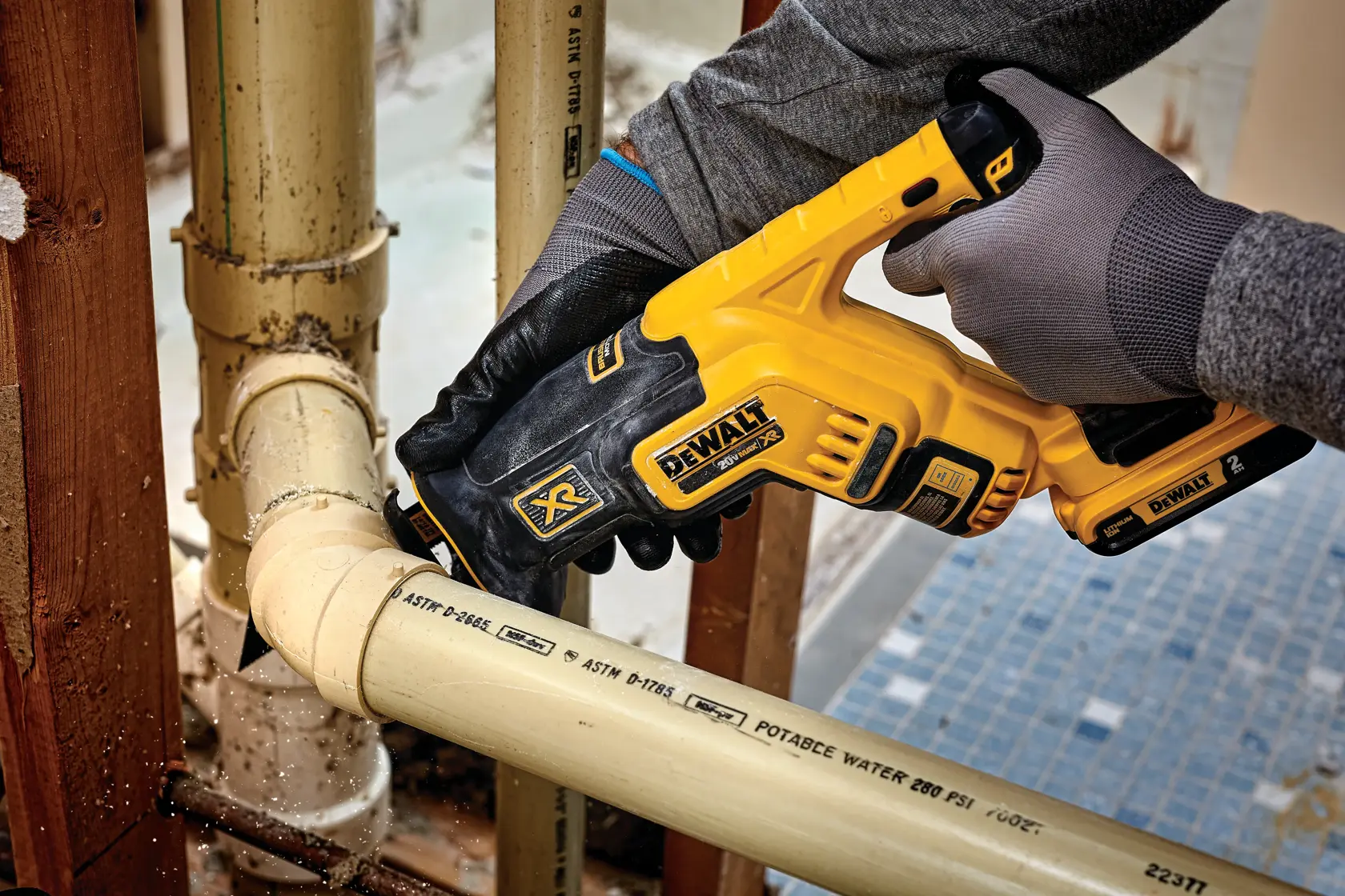 Close-up of a person using a DEWALT cordless reciprocating saw to cut a plastic pipe. The person is wearing gloves and working in a construction setting.