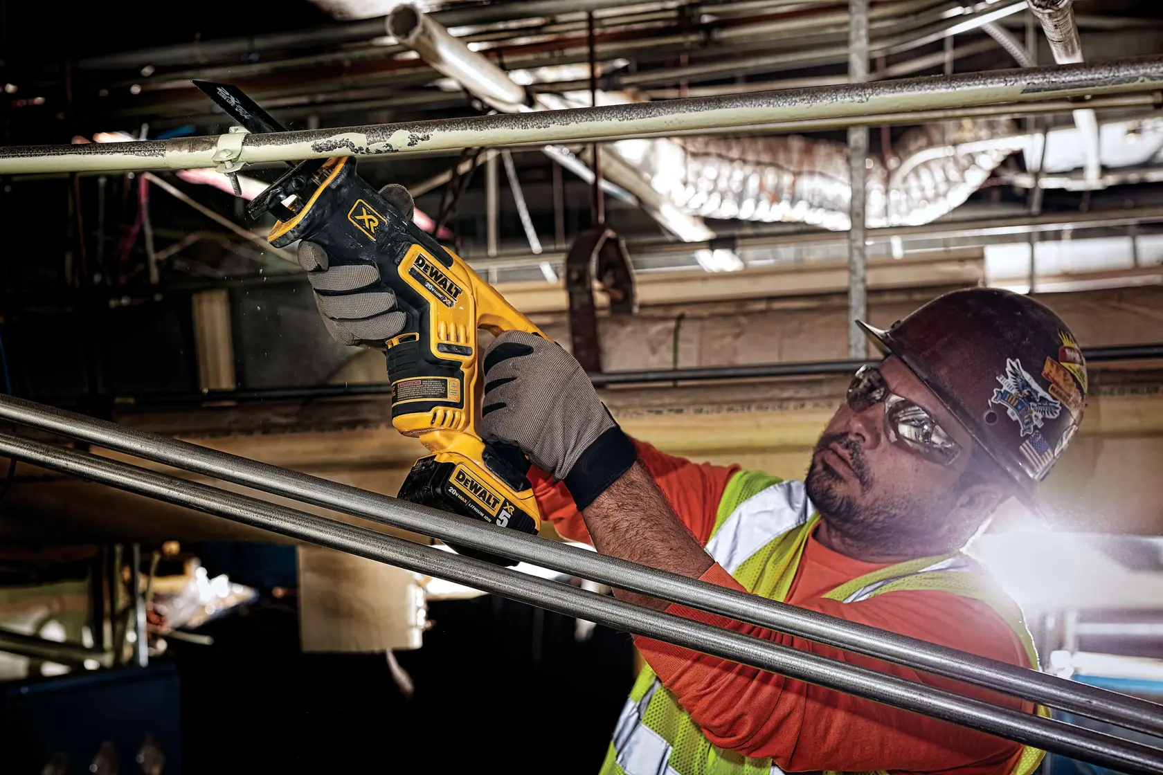 A worker using a DEWALT DCS367P1 cordless reciprocating saw to cut through a metal pipe, wearing gloves, safety vest, and helmet at an industrial work site.