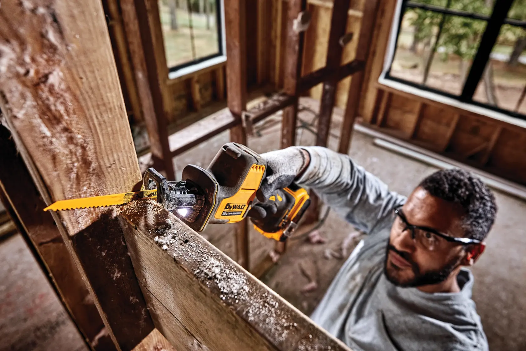 A person wearing gloves uses a DEWALT cordless reciprocating saw to cut through a wooden beam in a construction setting. The face of the person is obscured. The saw is yellow and black and features a visible blade, with sawdust present on the wood.