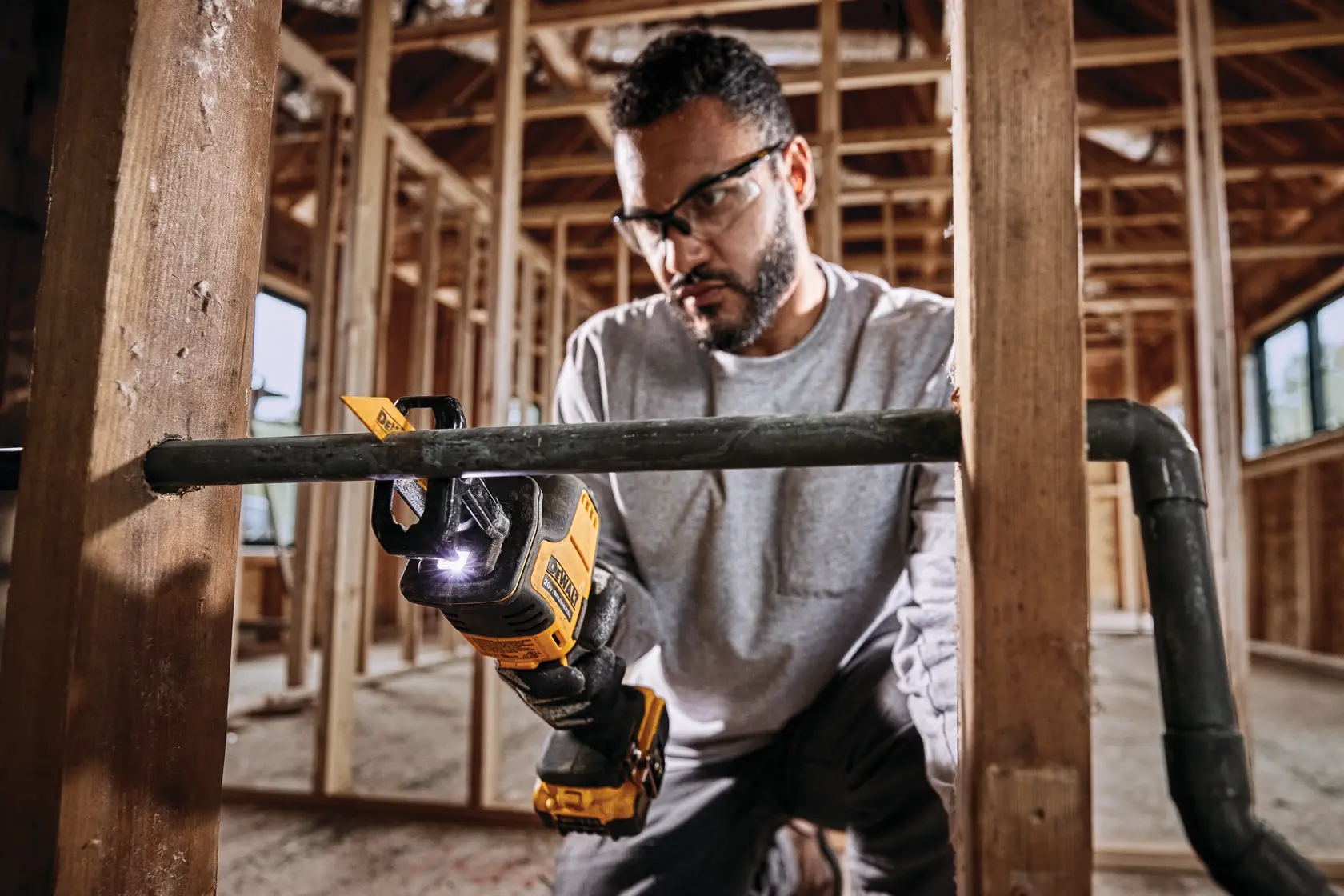 A person using a DEWALT cordless reciprocating saw to cut through a metal pipe in a construction setting, with safety gloves and wooden framing visible in the background. The person's face is blurred.
