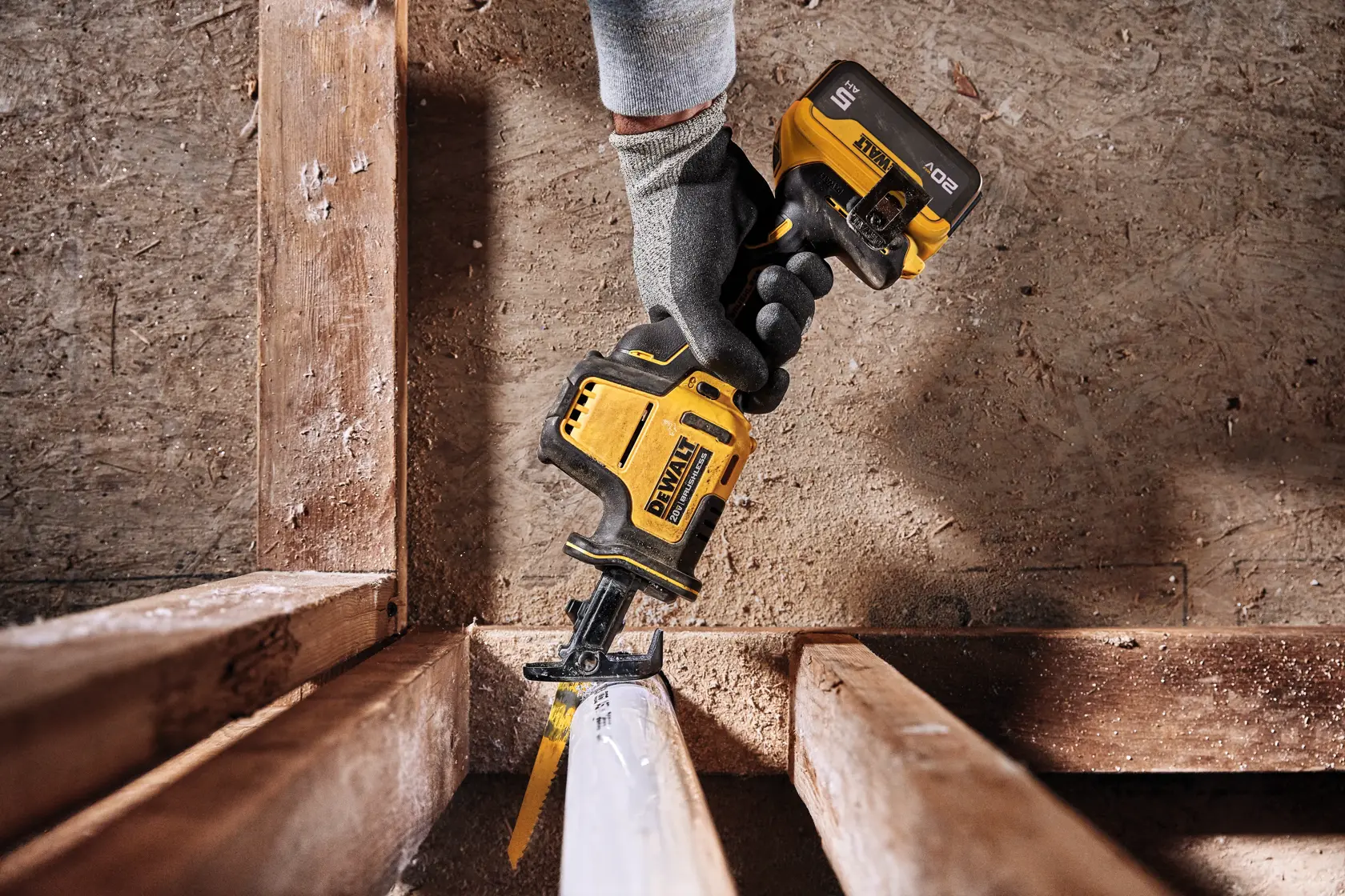 A gloved hand is using a yellow and black DEWALT cordless reciprocating saw to cut through a white plastic pipe in a wooden construction frame.