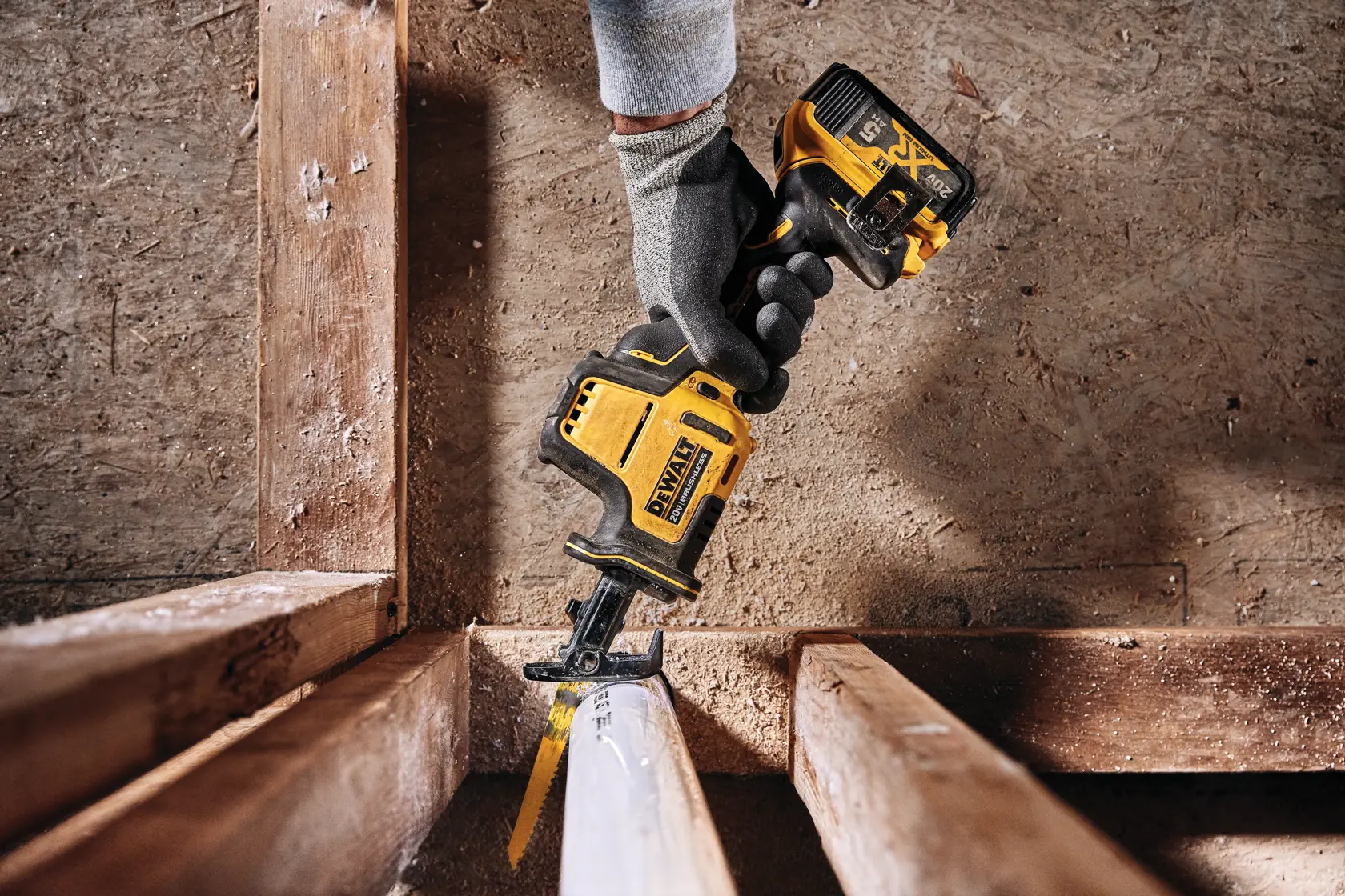 A person wearing gloves uses a DEWALT cordless reciprocating saw to cut through a white PVC pipe between wooden beams. The tool features a yellow and black design and is shown in a construction or renovation setting.