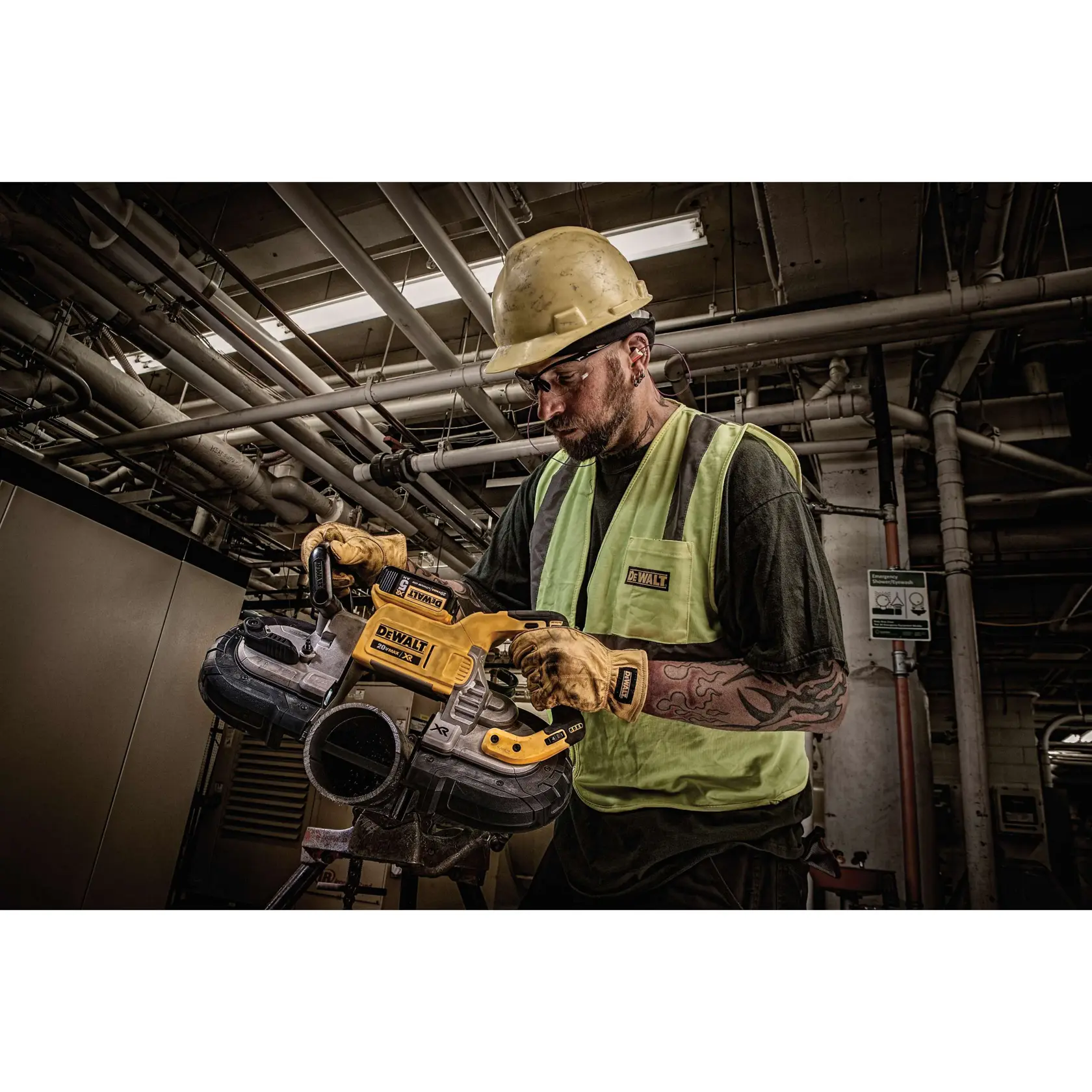 A person wearing safety gear, including a helmet, gloves, and a reflective vest, uses a DEWALT DCS374P2 cordless band saw in an industrial setting with visible pipes and machinery overhead. The person's face is blurred for privacy.