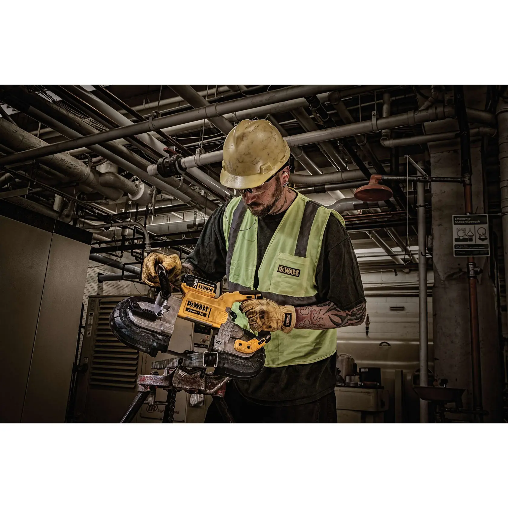 A worker wearing a yellow hard hat, gloves, and a reflective vest uses a DEWALT portable band saw to cut metal pipes in an industrial setting with complex piping overhead.