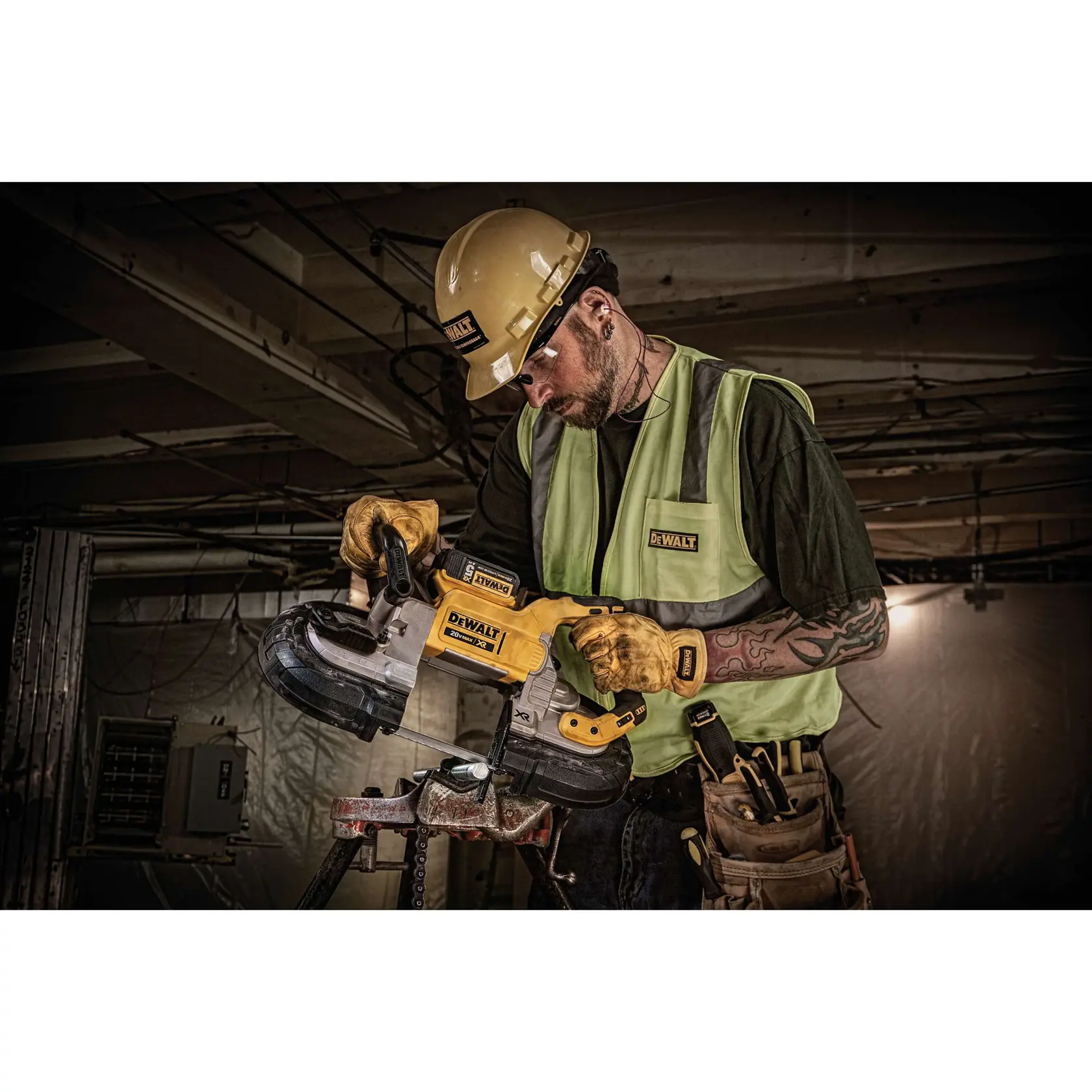 A construction worker wearing a DEWALT-branded helmet, safety vest, and gloves is using a DEWALT DCS374P2 portable band saw to cut metal on a worksite. The worker has tattoos on their arm and is equipped with a tool belt, working in an industrial environment.