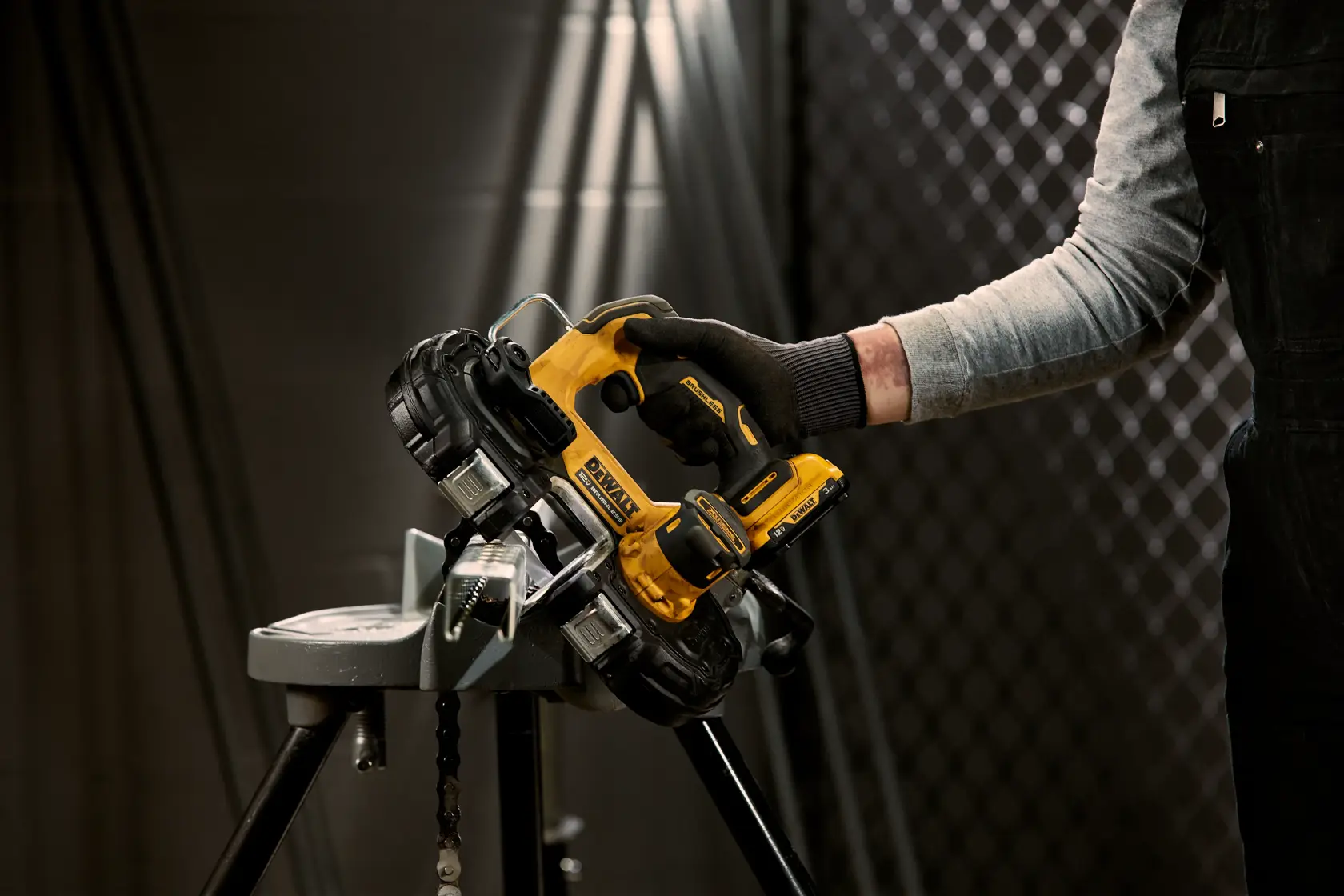 A person wearing work gloves is holding a yellow DEWALT portable band saw, model DCS375B, and cutting a metal piece in an industrial setting.