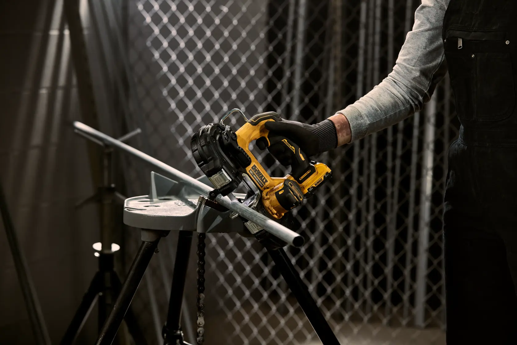 A person using a yellow and black DEWALT portable band saw to cut a metal pipe. The pipe is secured on a metal stand in an industrial setting with a chain-link fence in the background.