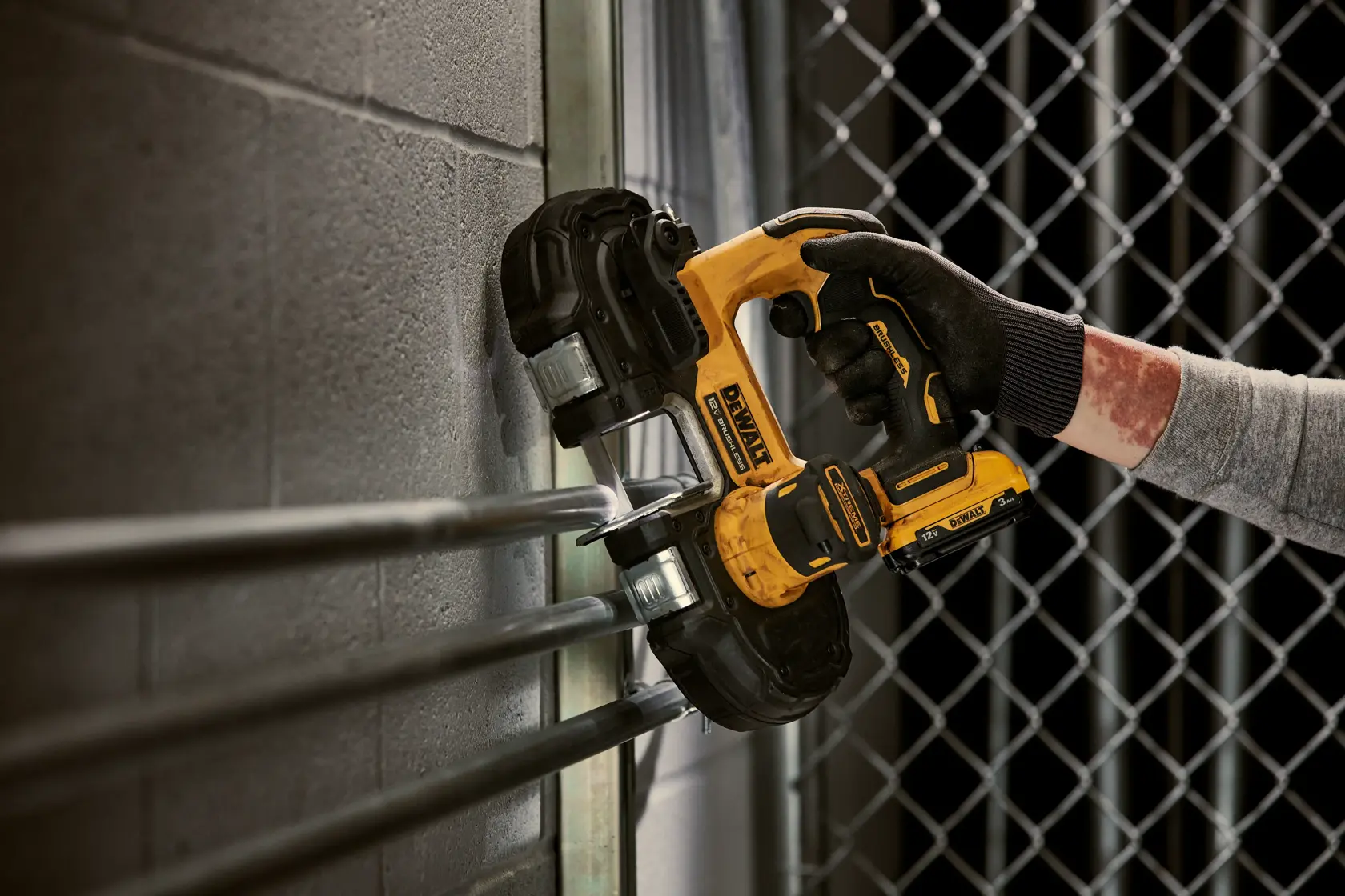 A person wearing gloves is using a DEWALT DCS375B cordless band saw to cut metal pipes mounted on a wall near a chain-link fence.