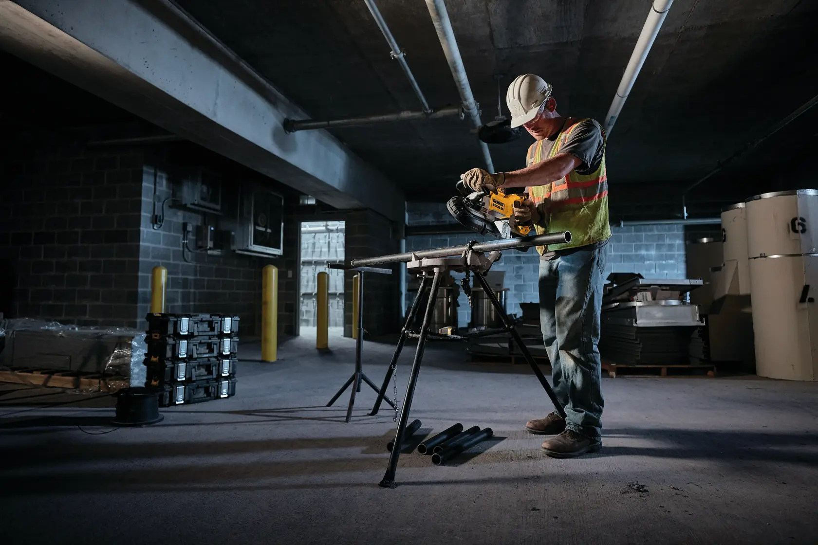 A construction worker wearing a hard hat and safety vest uses a DEWALT power tool to cut metal pipes in an industrial setting, standing at a workbench with equipment and materials nearby.