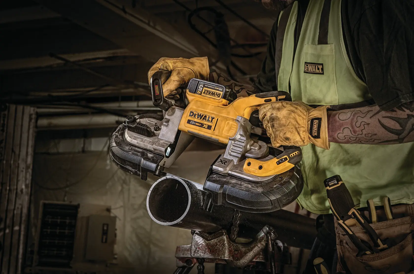 A person wearing work gloves uses a DEWALT cordless band saw to cut a metal pipe in an industrial setting. The DEWALT logo is visible on the tool, gloves, and shirt.