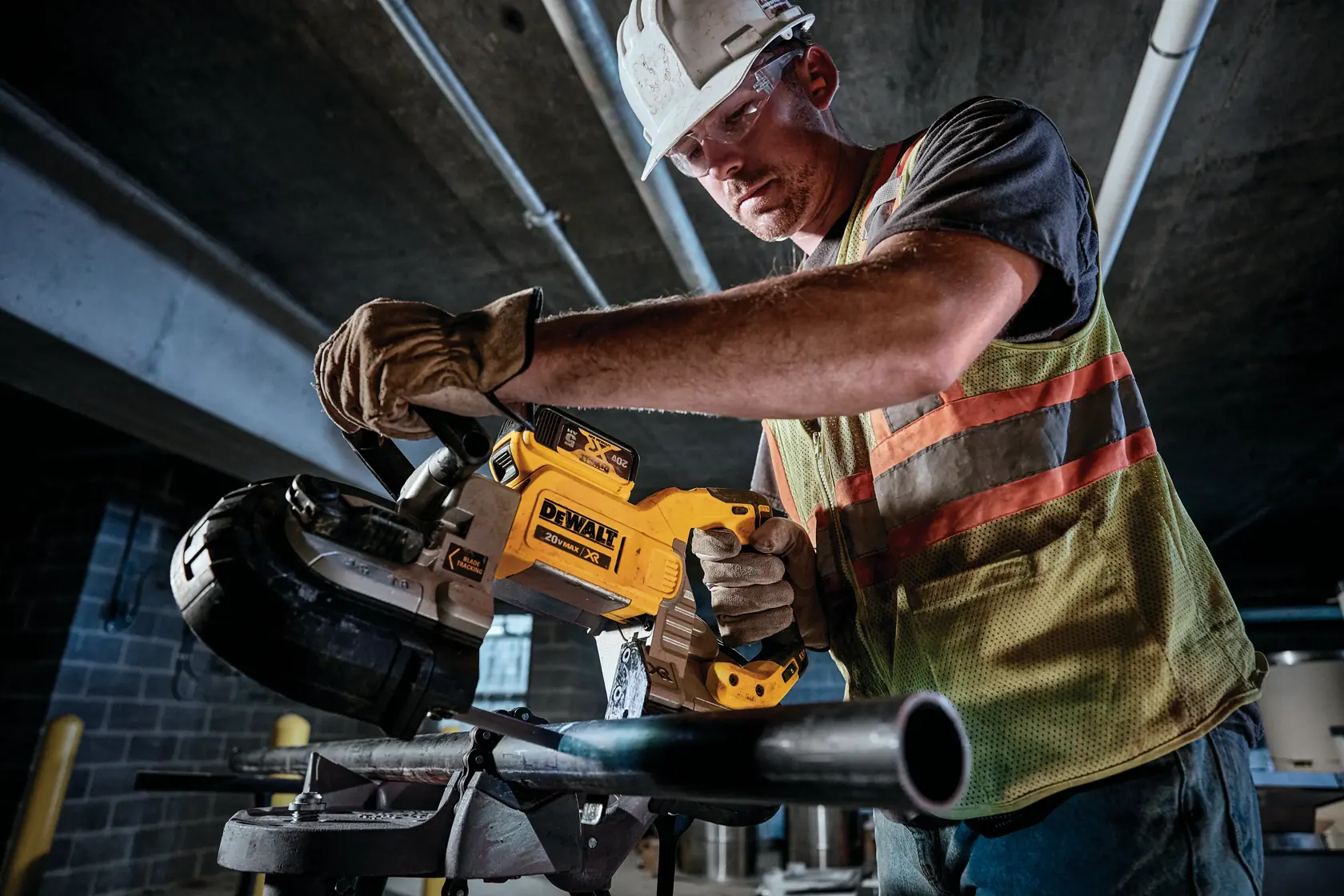A construction worker wearing safety gear is using a DEWALT DCS376P2 cordless band saw to cut a metal pipe in an industrial setting.