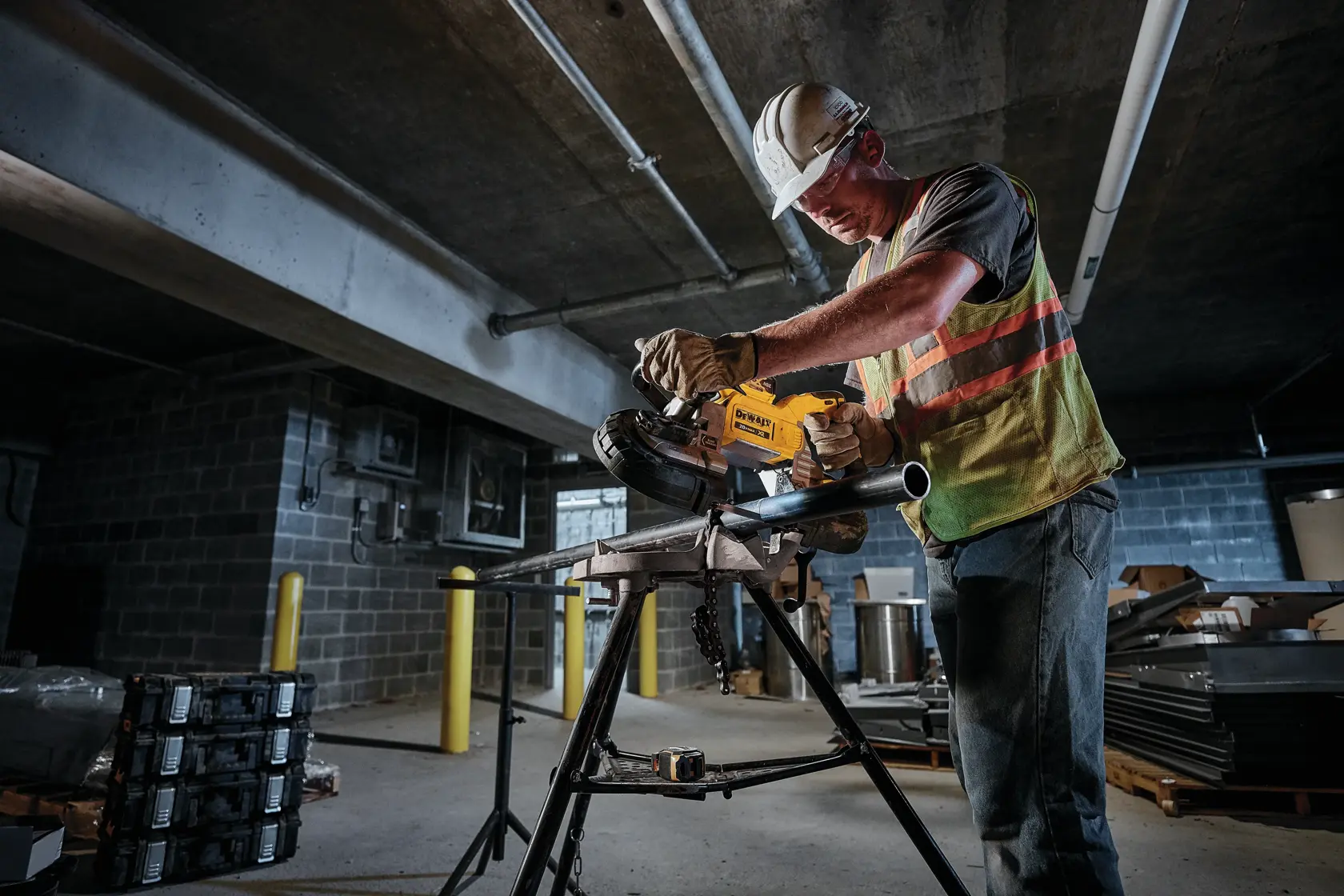 A construction worker wearing a safety vest and helmet is using a DEWALT power tool to cut a metal pipe on a work stand in an industrial indoor setting.