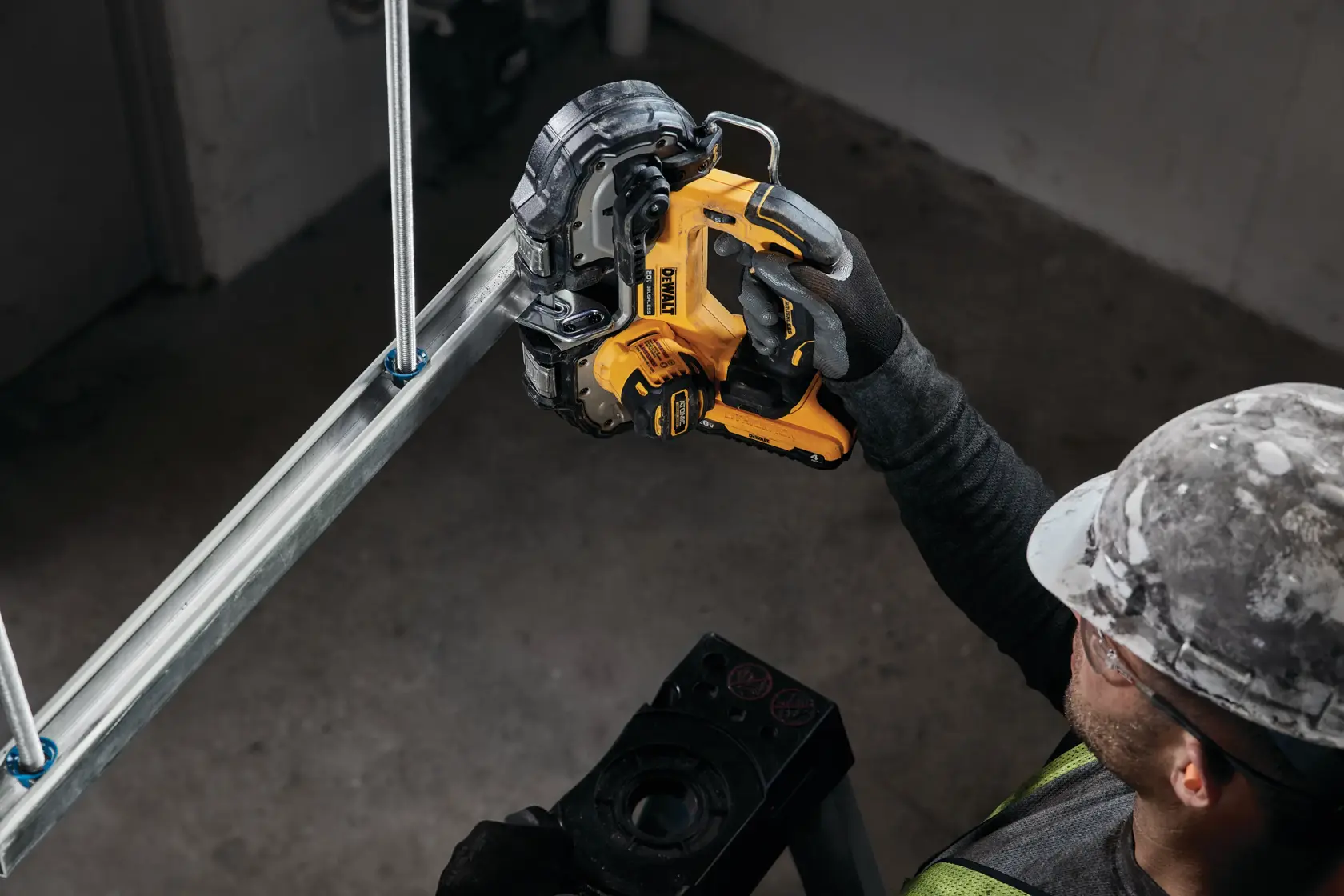 A person wearing a safety helmet and gloves uses a DEWALT cordless band saw to cut a metal pipe indoors.