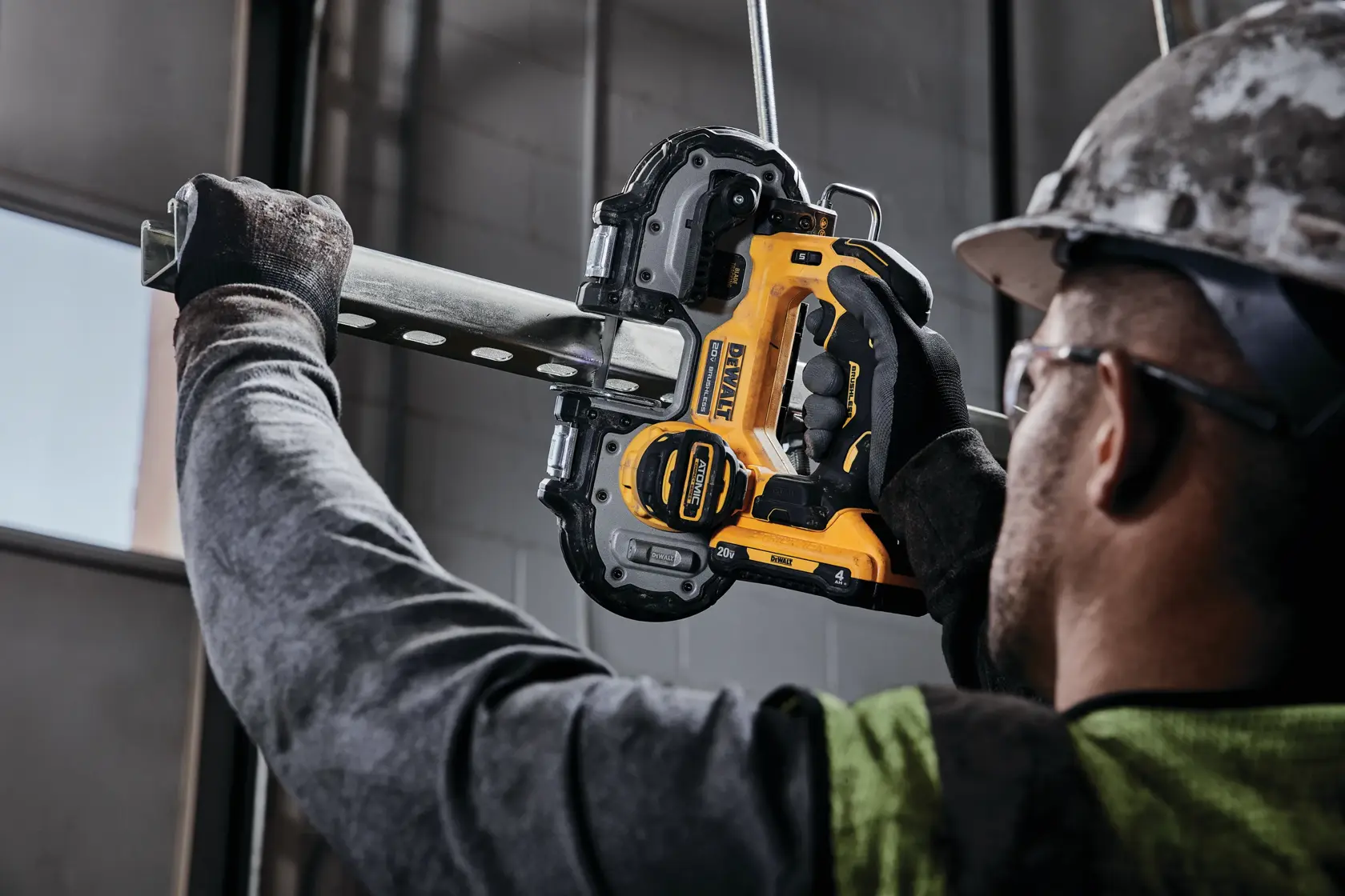 A person wearing gloves and a hard hat uses a DEWALT cordless band saw to cut a metal pipe indoors.