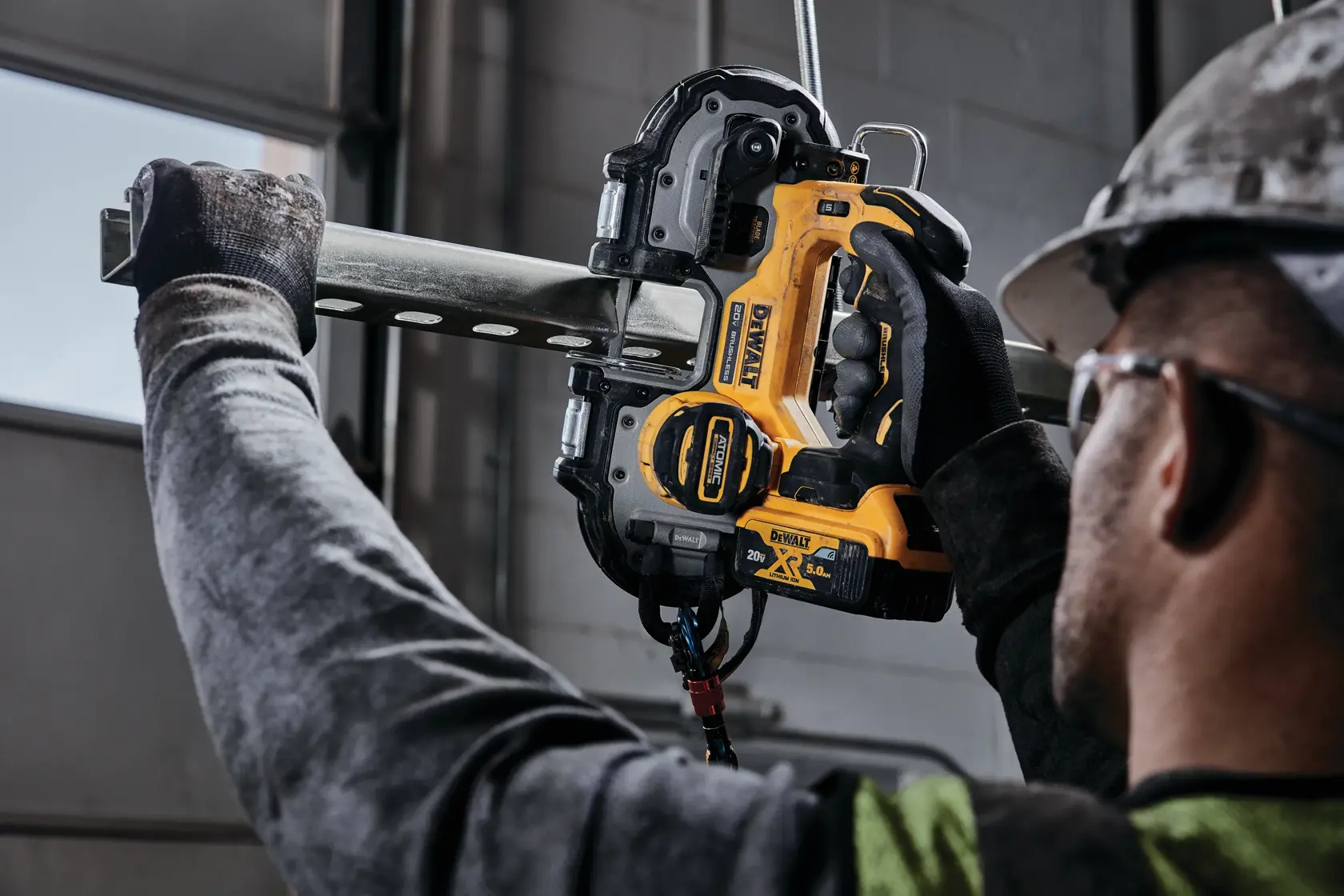 A worker using a DEWALT DCS377B 20V XR cordless band saw to cut a metal pipe indoors. The worker is wearing gloves, a safety vest, and a protective helmet.