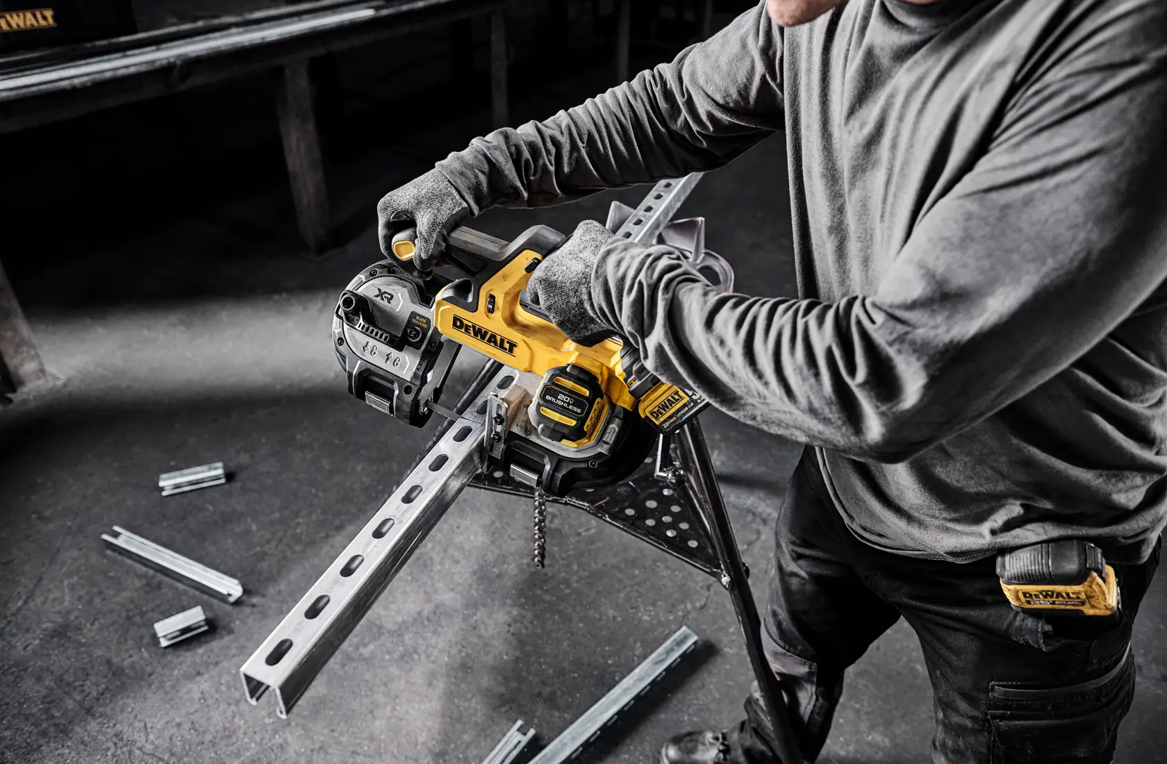 A person using a DEWALT cordless band saw to cut a metal channel on a workbench. The person is wearing gloves and work clothes, with several metal pieces and tools visible in the background.