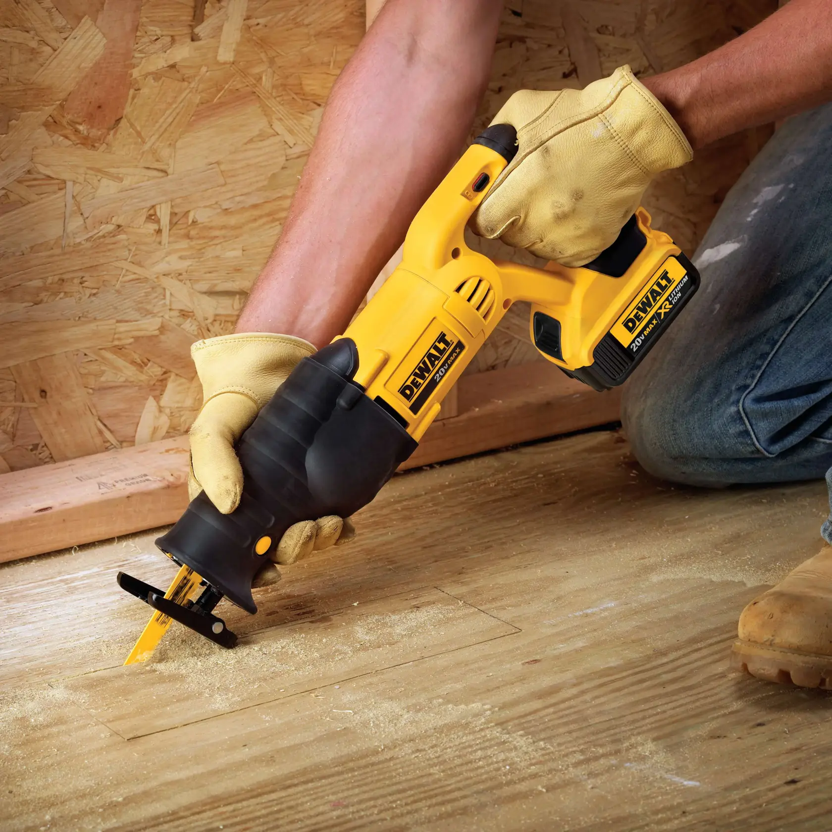 A person wearing work gloves uses a DEWALT cordless reciprocating saw to cut into a wooden floor. The saw is yellow and black, and sawdust is visible around the cutting area.