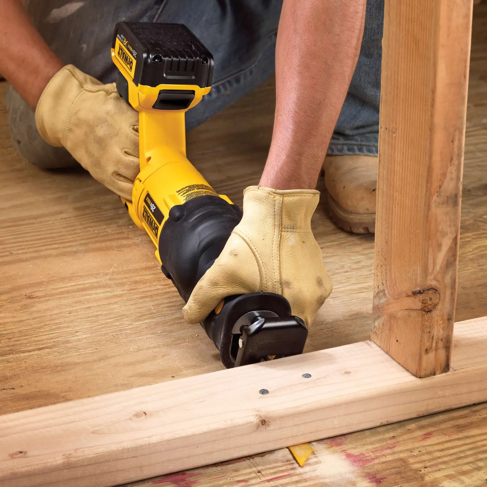 A person wearing yellow work gloves is using a yellow DEWALT cordless reciprocating saw to cut wood on a construction site. The saw is positioned close to a wooden support beam and a floor made of wooden planks.