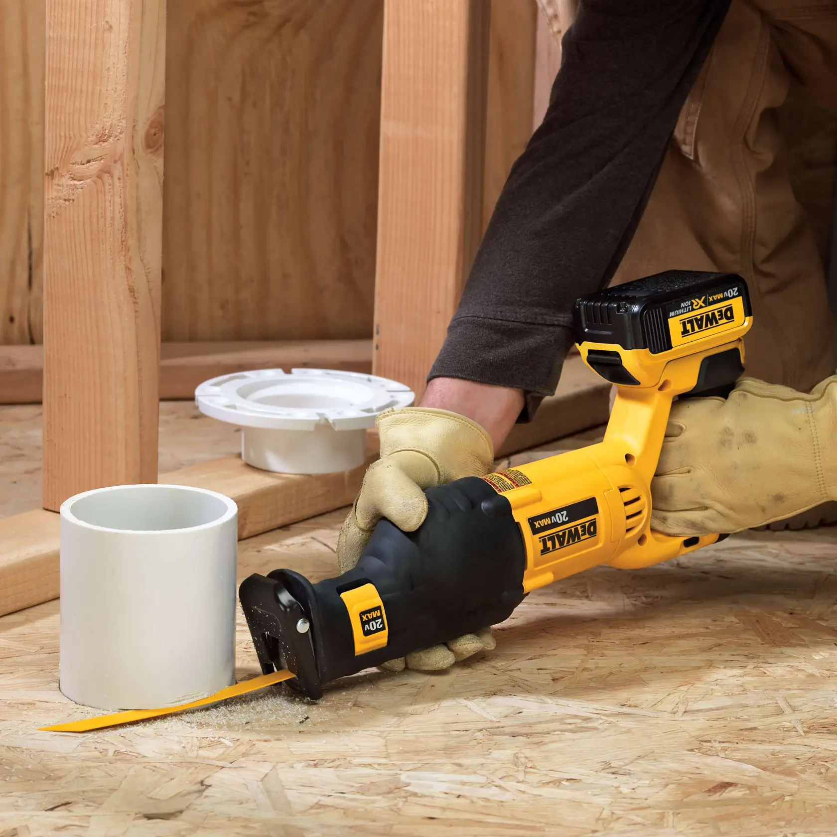 A person wearing work gloves uses a DEWALT 20V Max cordless reciprocating saw to cut a white PVC pipe on a wooden floor, with wooden framing and plumbing components visible in the background.