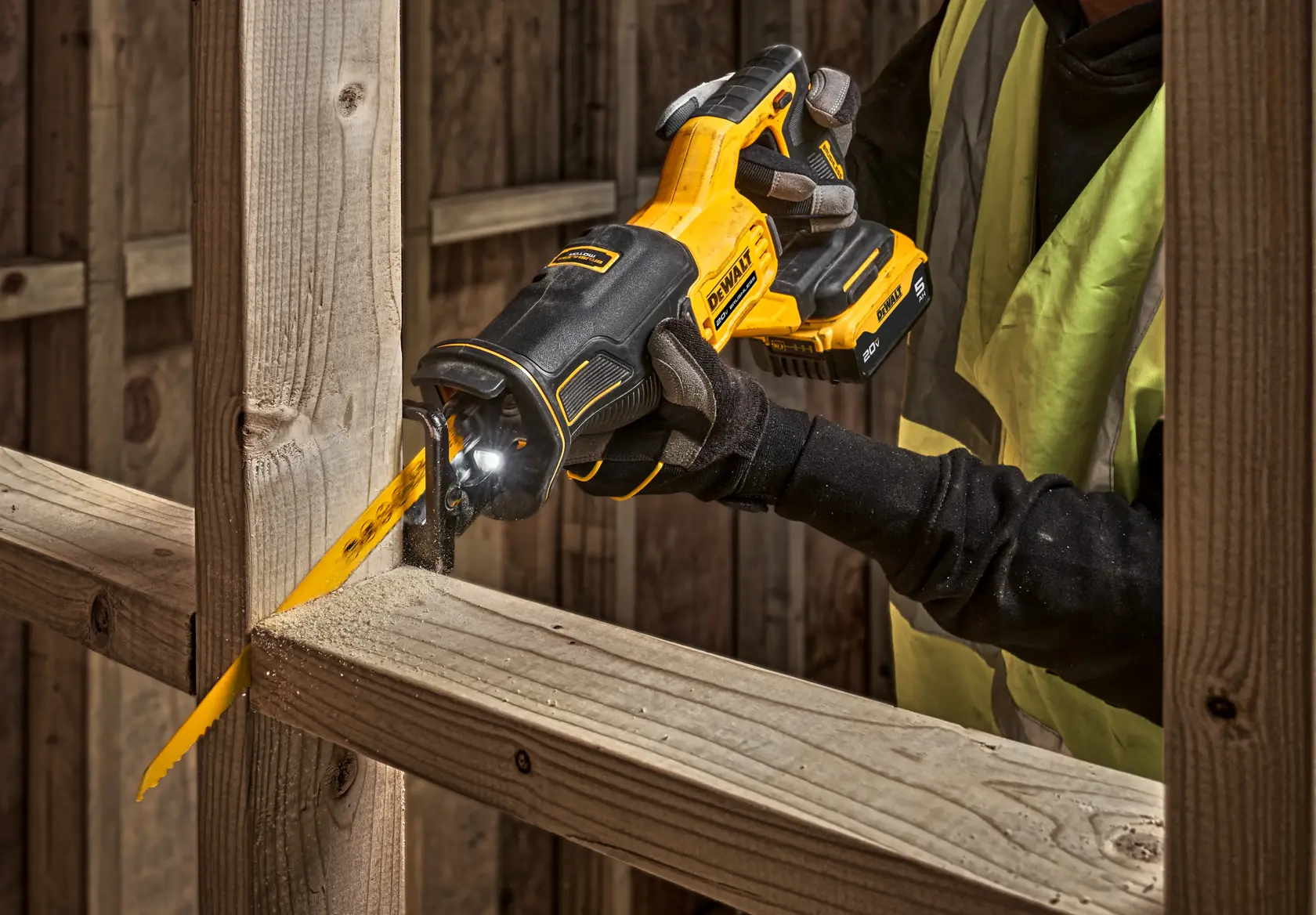 A person wearing gloves and a safety vest uses a DEWALT cordless reciprocating saw to cut through a wooden beam at a construction site.