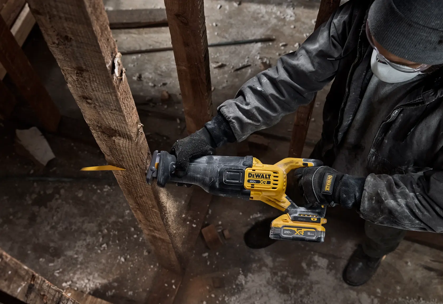 A person wearing gloves, a mask, and a beanie is using a DEWALT cordless reciprocating saw to cut through a wooden beam inside a construction or renovation site.