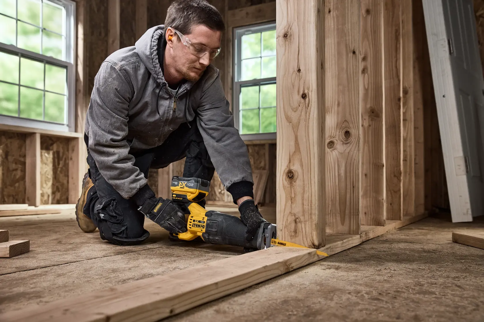 A person wearing work gloves and ear protection uses a DEWALT reciprocating saw to cut wooden boards inside a room under construction.