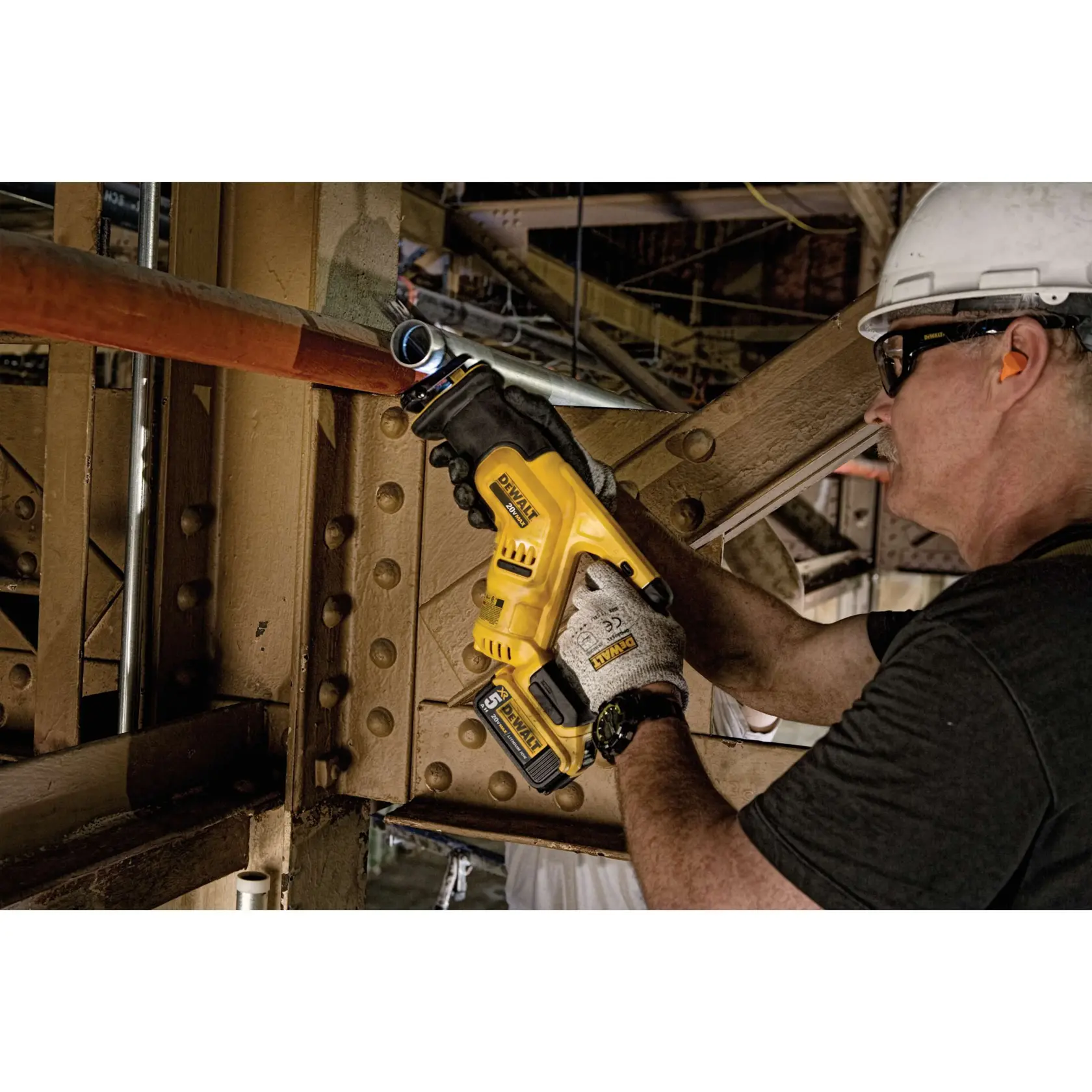 A person wearing gloves and a hard hat uses a DEWALT DCS387B cordless reciprocating saw to cut through a metal pipe in an industrial setting.