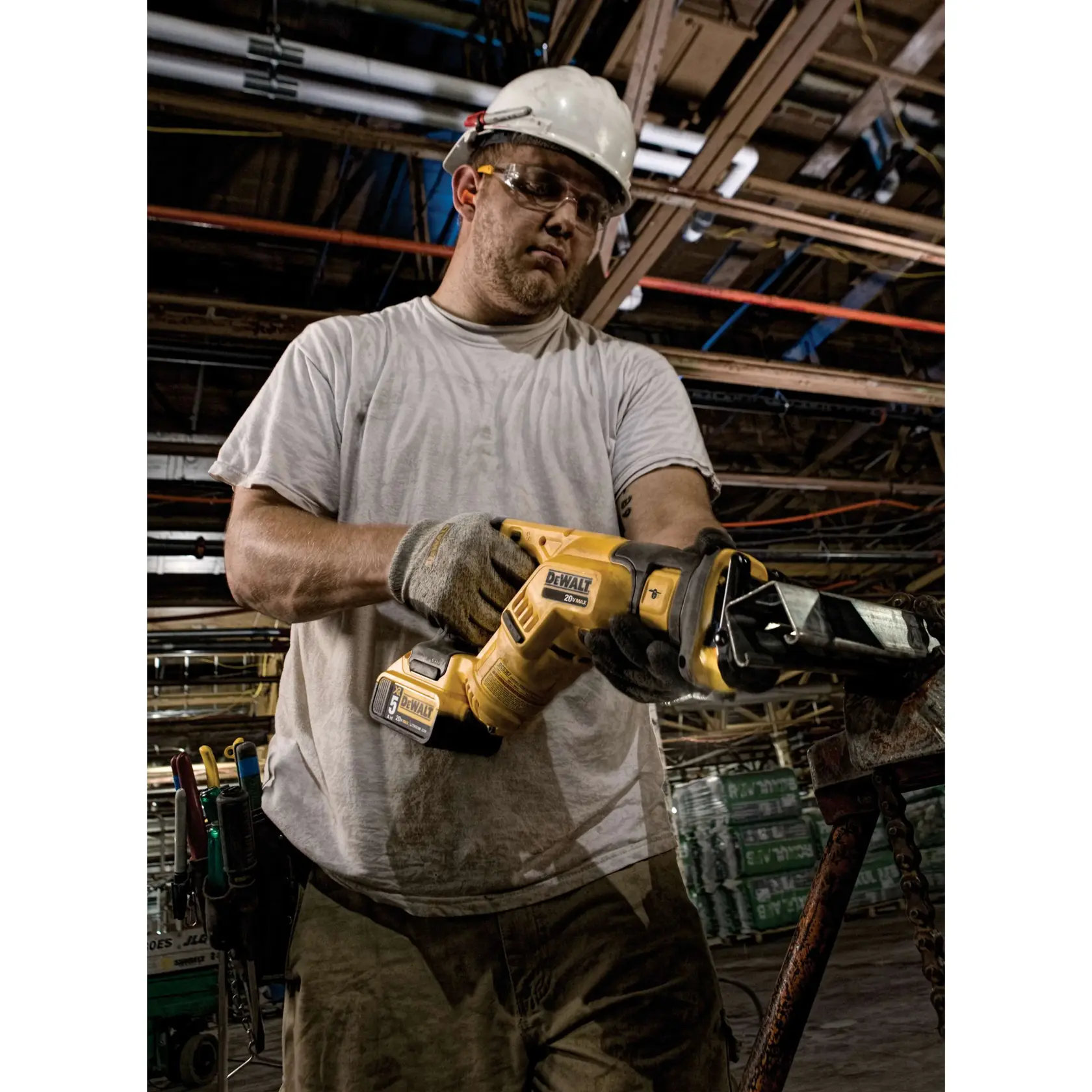 A person wearing work gloves and a hard hat is operating a DEWALT DCS387B reciprocating saw in an industrial setting. The background shows construction materials and exposed ceiling pipes.