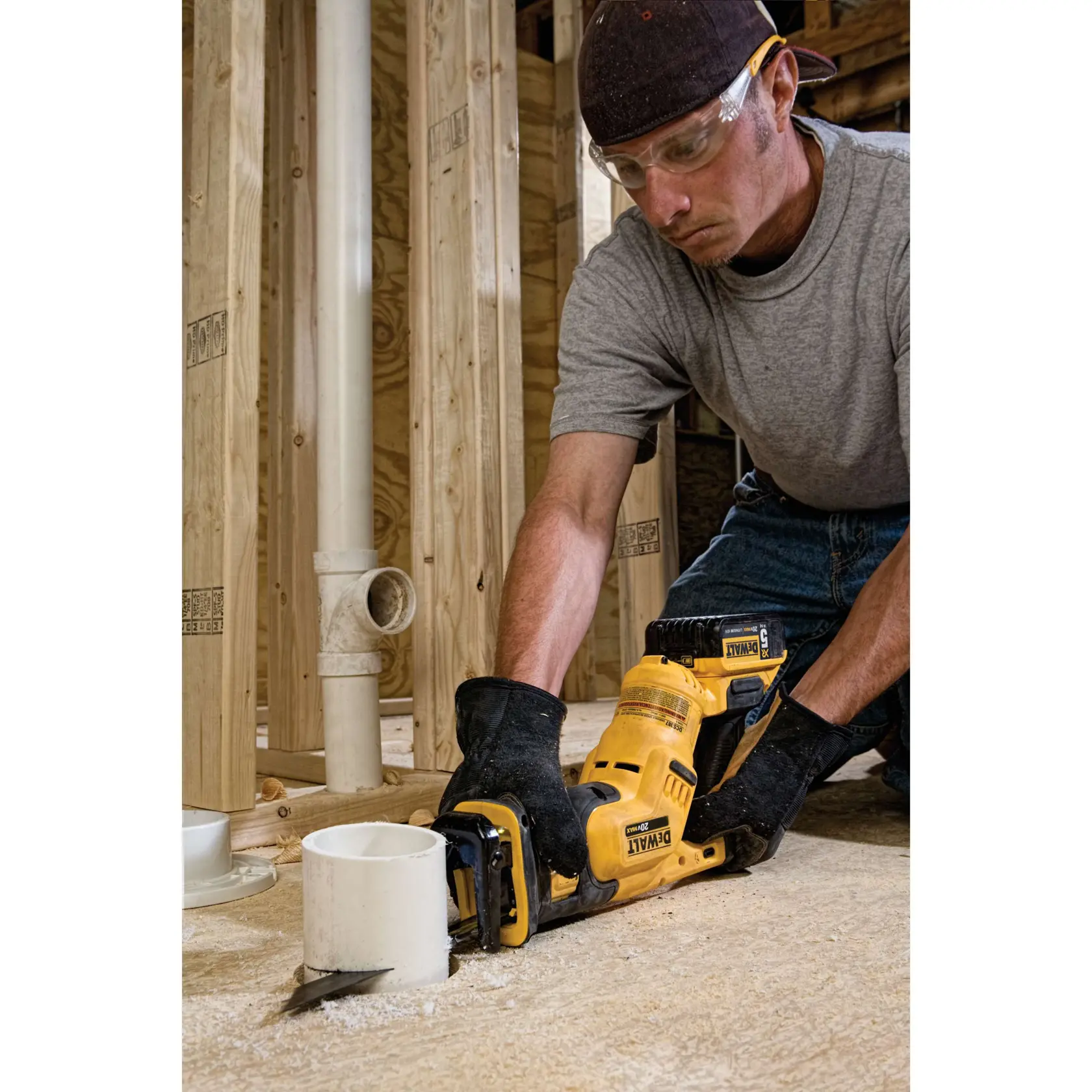 A person wearing gloves and a cap is using a DEWALT cordless reciprocating saw to cut a PVC pipe on a construction site with wooden framing and exposed plumbing.
