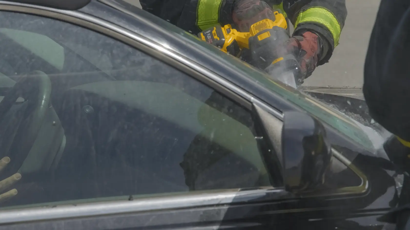A person wearing safety gloves and a high-visibility jacket is using a DEWALT DCS389B power tool to cut through the window frame of a black car.