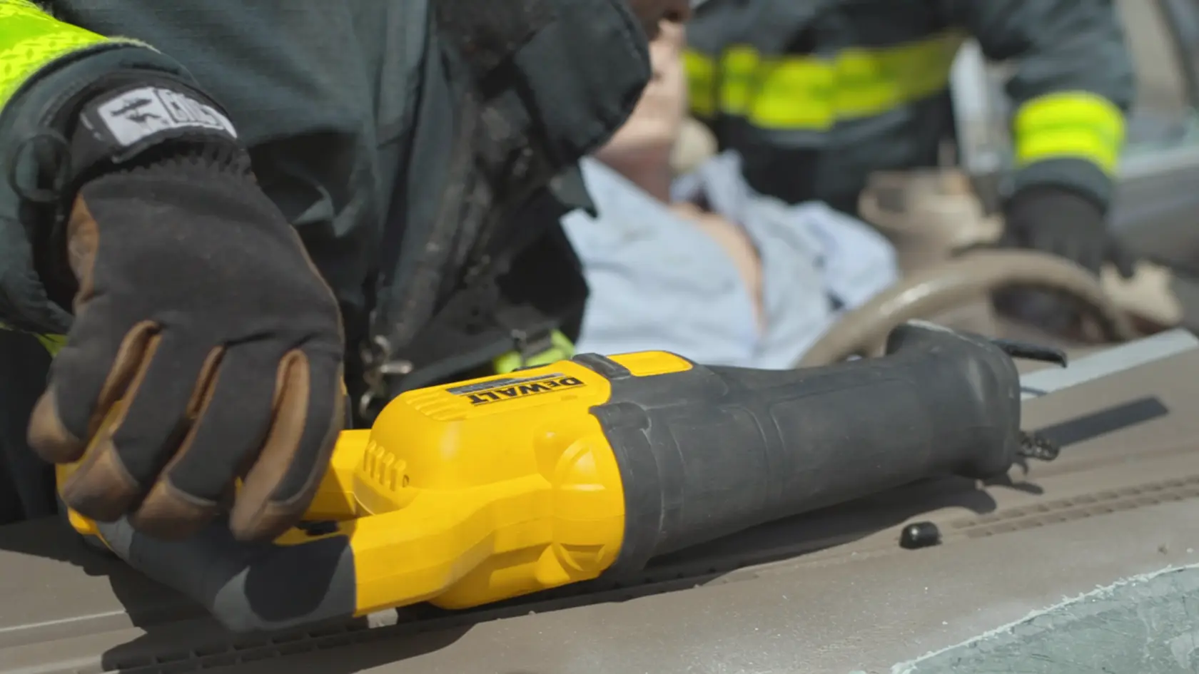 A close-up of a DEWALT DCS389B reciprocating saw being used by a person wearing gloves and protective clothing, with a blurred background showing another individual and a vehicle interior.