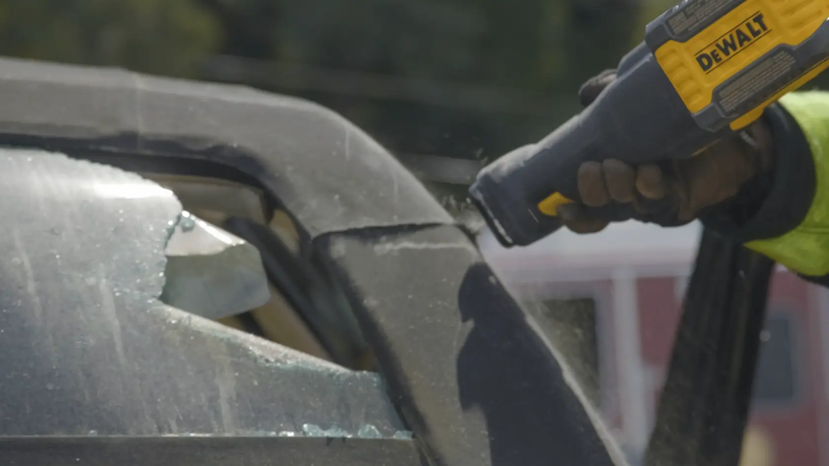 A close-up of a person using a DEWALT cordless reciprocating saw to cut through the frame of a vehicle near a broken window.