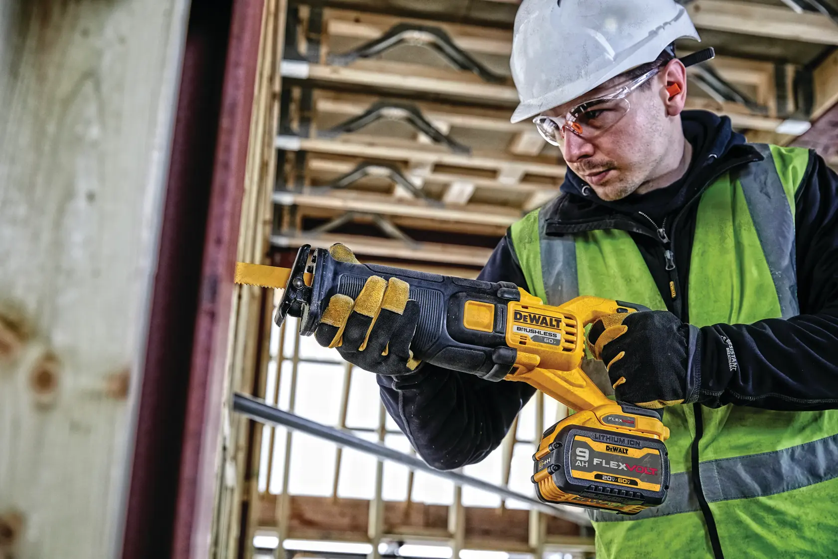 A person wearing a hard hat and safety vest uses a DEWALT DCS389X1 cordless reciprocating saw with a FLEXVOLT battery at a construction site. The person's face is blurred for privacy.