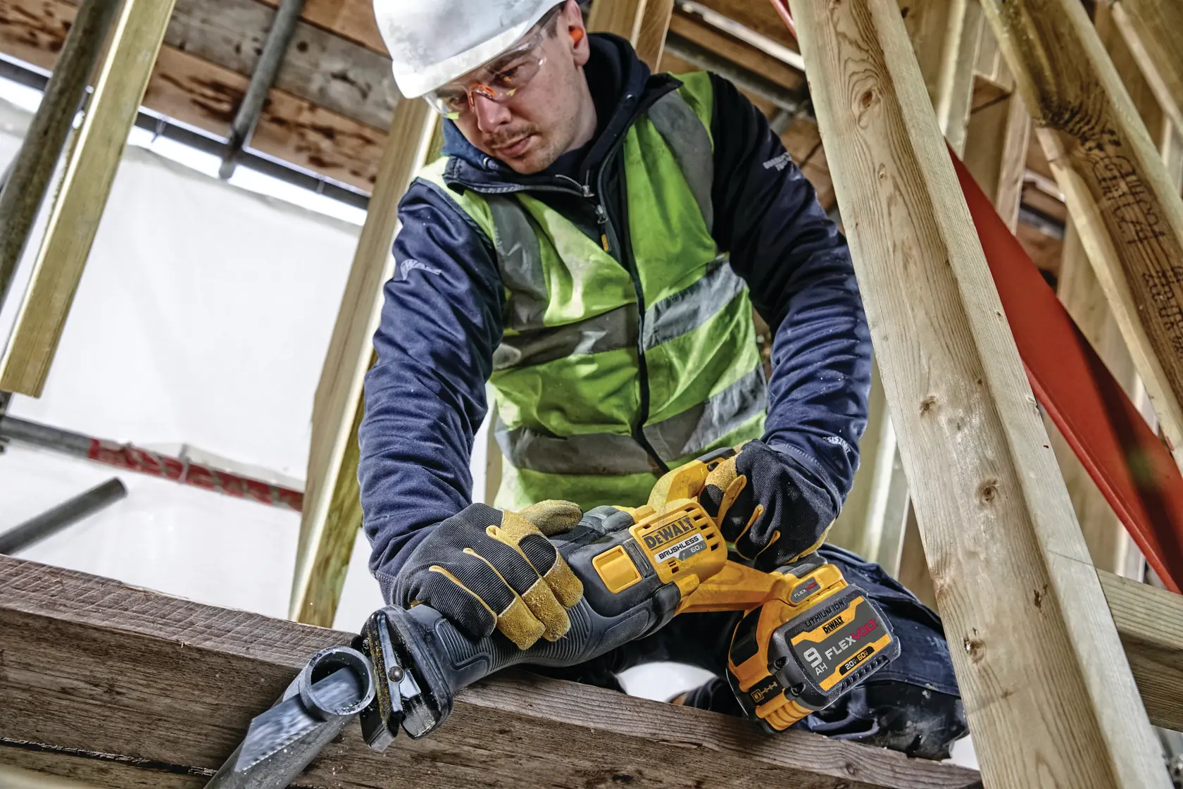 A construction worker using a DEWALT cordless reciprocating saw (SKU: DCS389X1) with FlexVolt battery to cut wood beams at a construction site. The worker is wearing safety gloves, a high-visibility vest, and a hard hat.