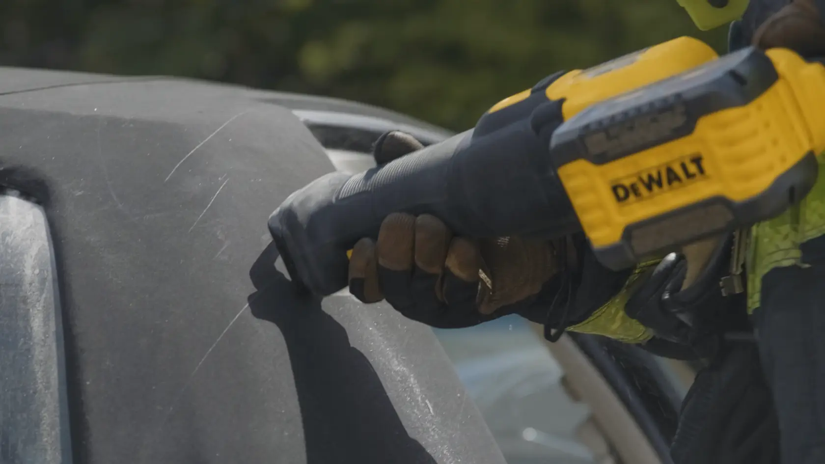 A person wearing gloves is using a DEWALT cordless reciprocating saw to cut through a dark surface, possibly part of a vehicle.