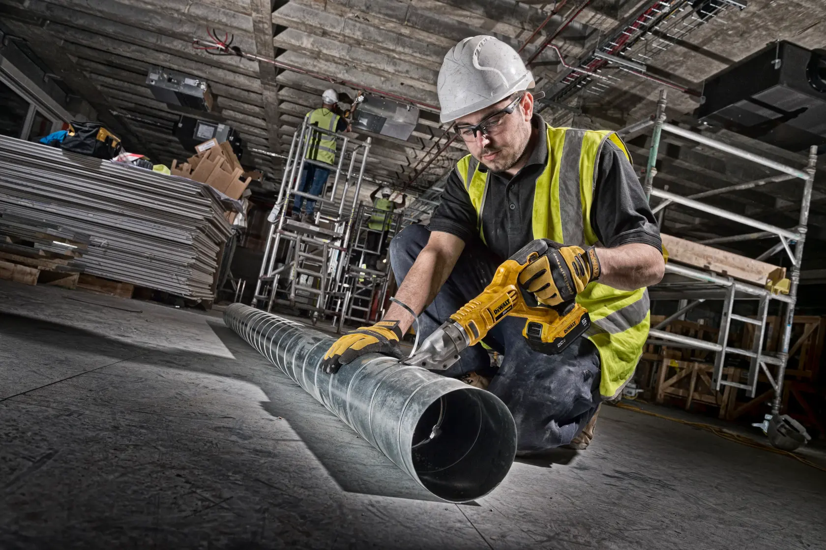 A construction worker wearing a hard hat and high-visibility vest is using a yellow power tool to cut a metal ventilation duct on the floor of an industrial site. In the background, another worker stands on scaffolding with piles of construction materials nearby.