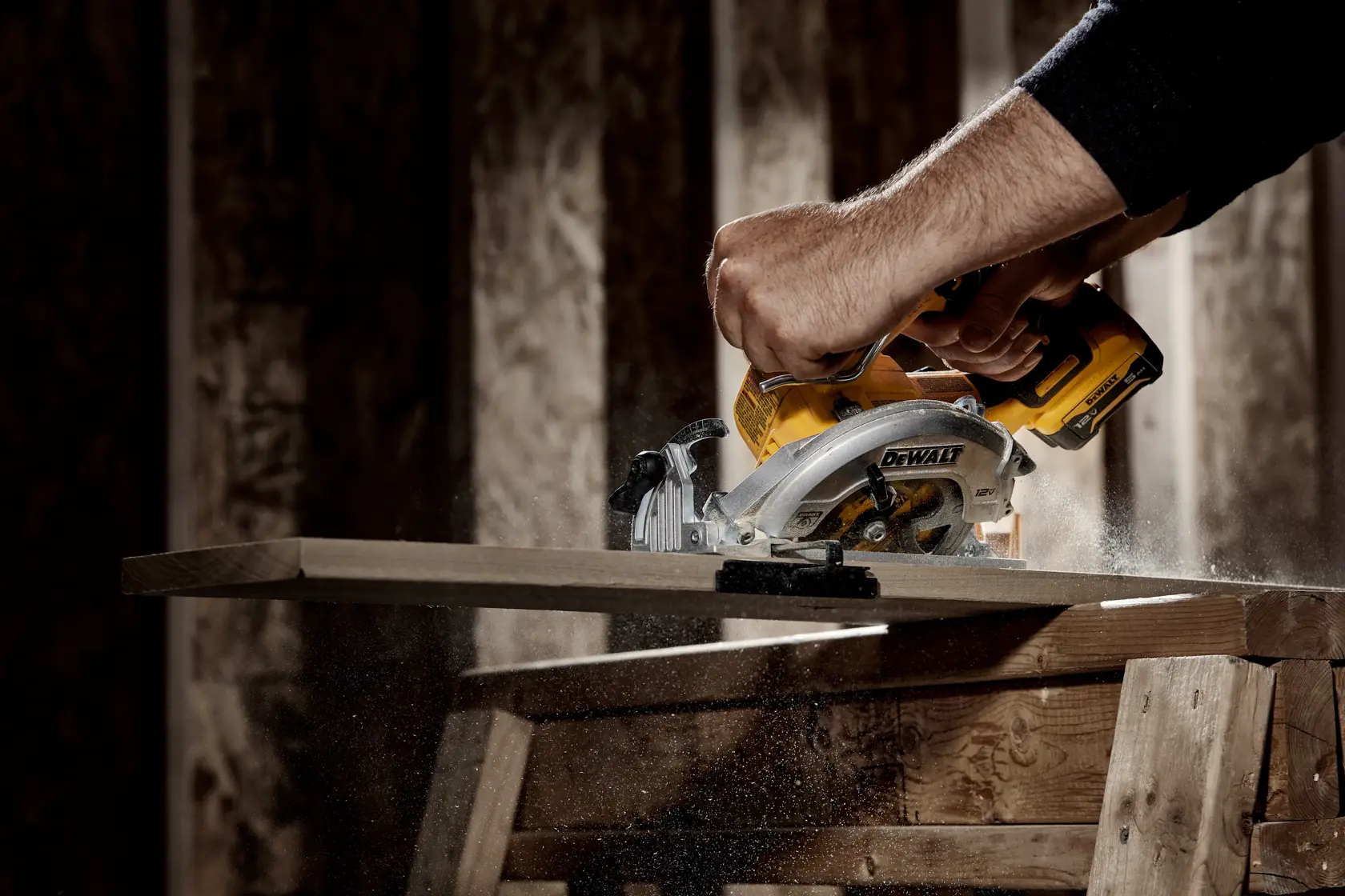 A person using a DEWALT cordless circular saw to cut a wooden plank, with sawdust flying in a workshop setting.