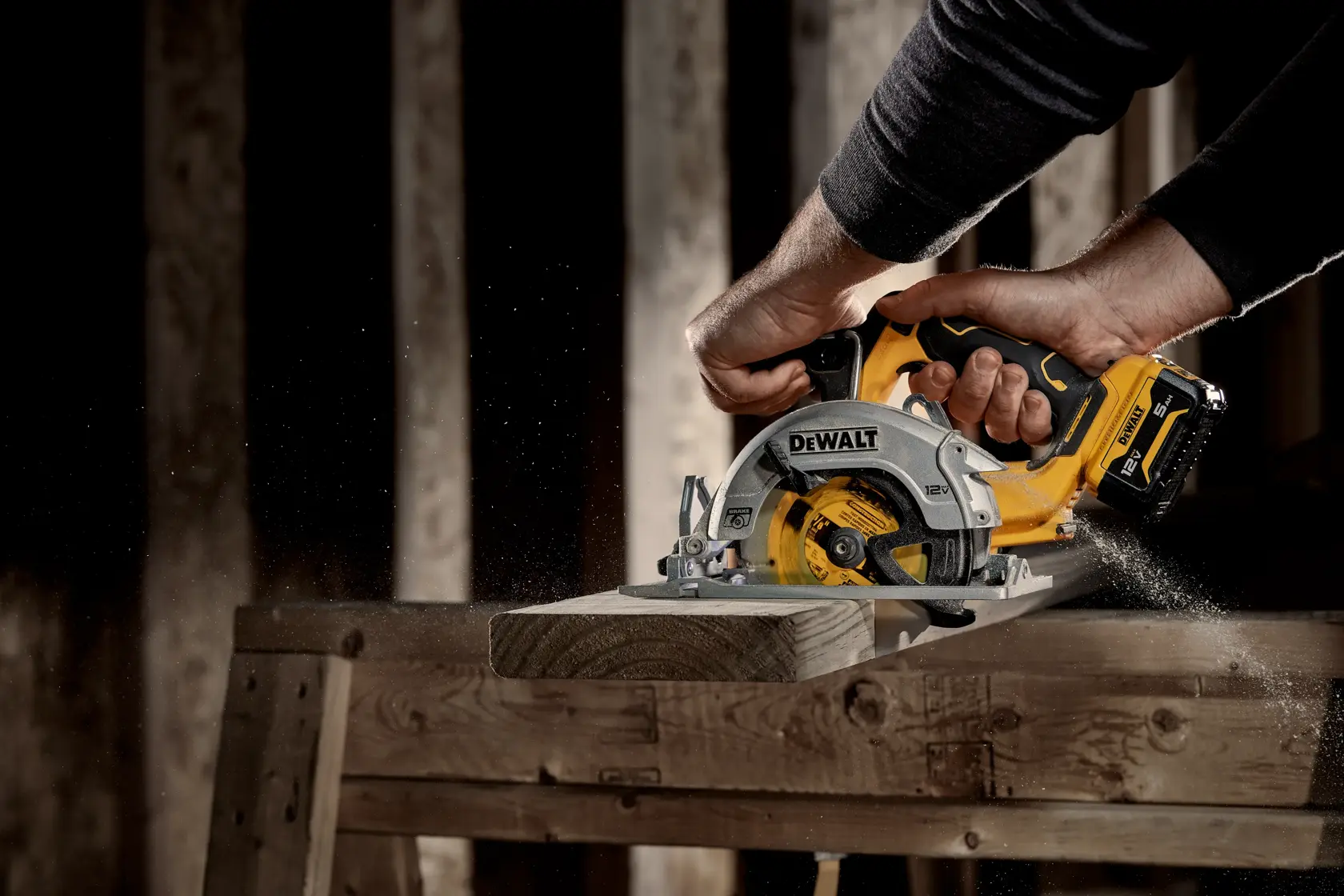 A DEWALT cordless circular saw in use, cutting through a piece of wood on a workbench. Wood shavings are visible as the saw operates, and the tool is being held by a person wearing a dark long-sleeve shirt.