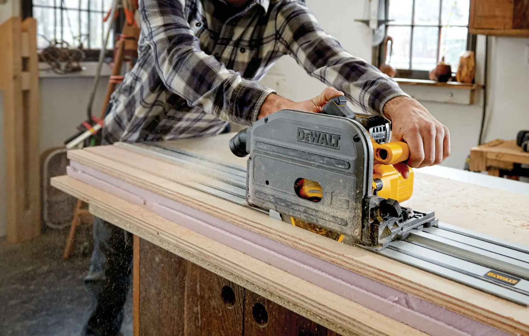 A person operating a DEWALT DCS520B track saw to cut a sheet of wood on a workbench in a workshop. The saw is mounted on a guide rail, and sawdust is visible as the person pushes the tool.