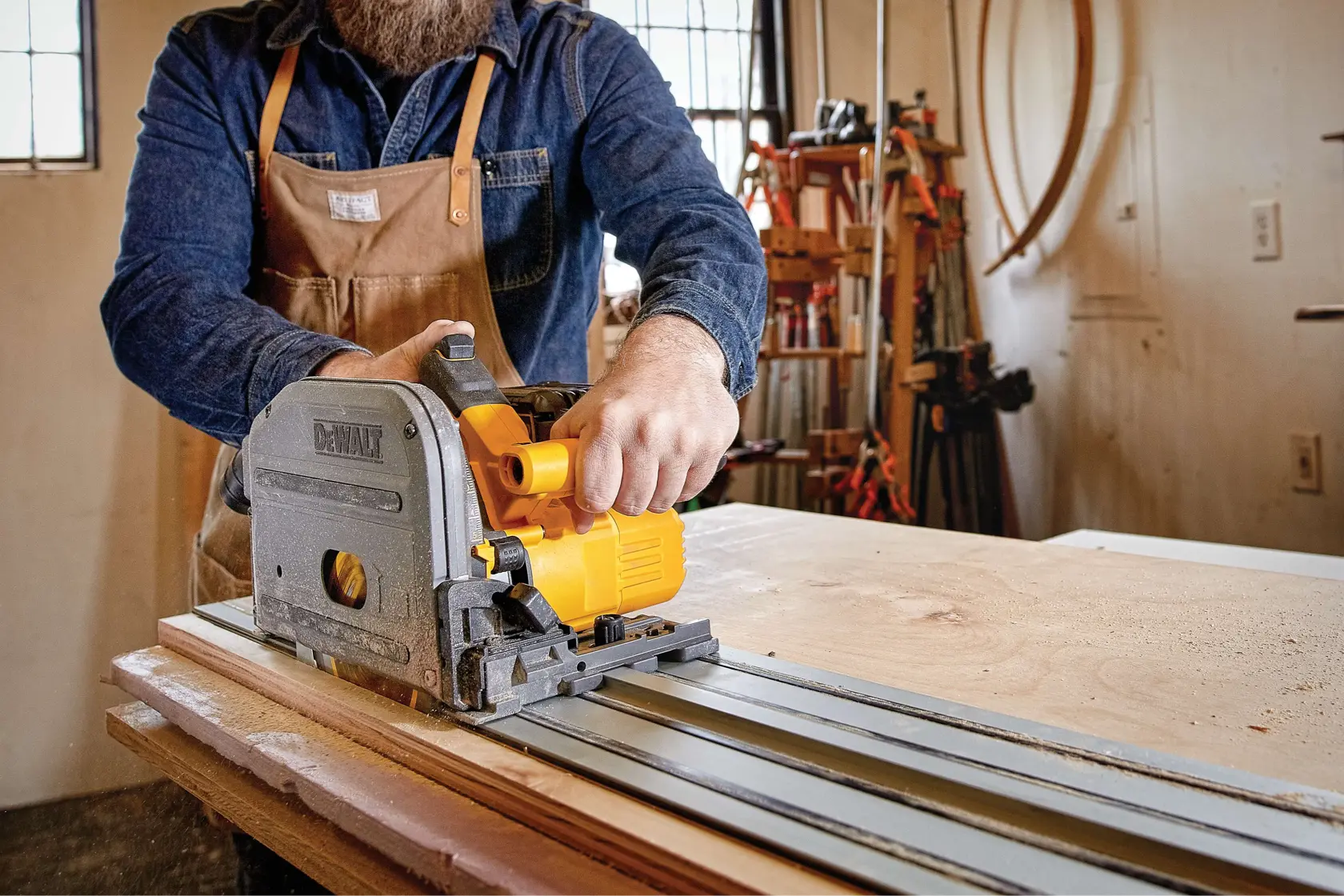 A person wearing a denim shirt and brown apron operates a DEWALT track saw to cut a sheet of wood in a workshop.