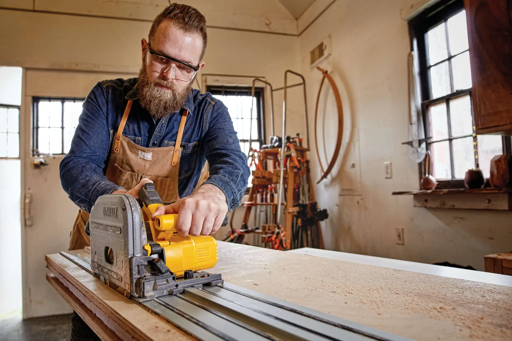 A person wearing a denim shirt and apron operates a DEWALT track saw cutting wood on a workbench in a workshop. The person's face is blurred.