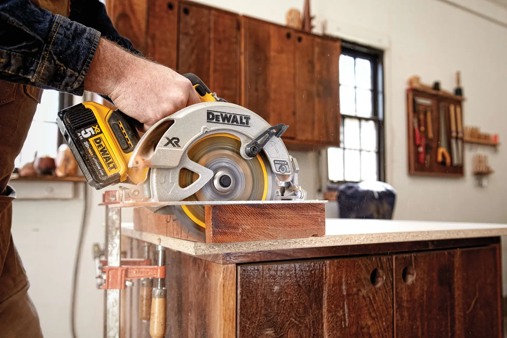 A person using a DEWALT cordless circular saw to cut a piece of wood on a workbench in a woodworking shop.
