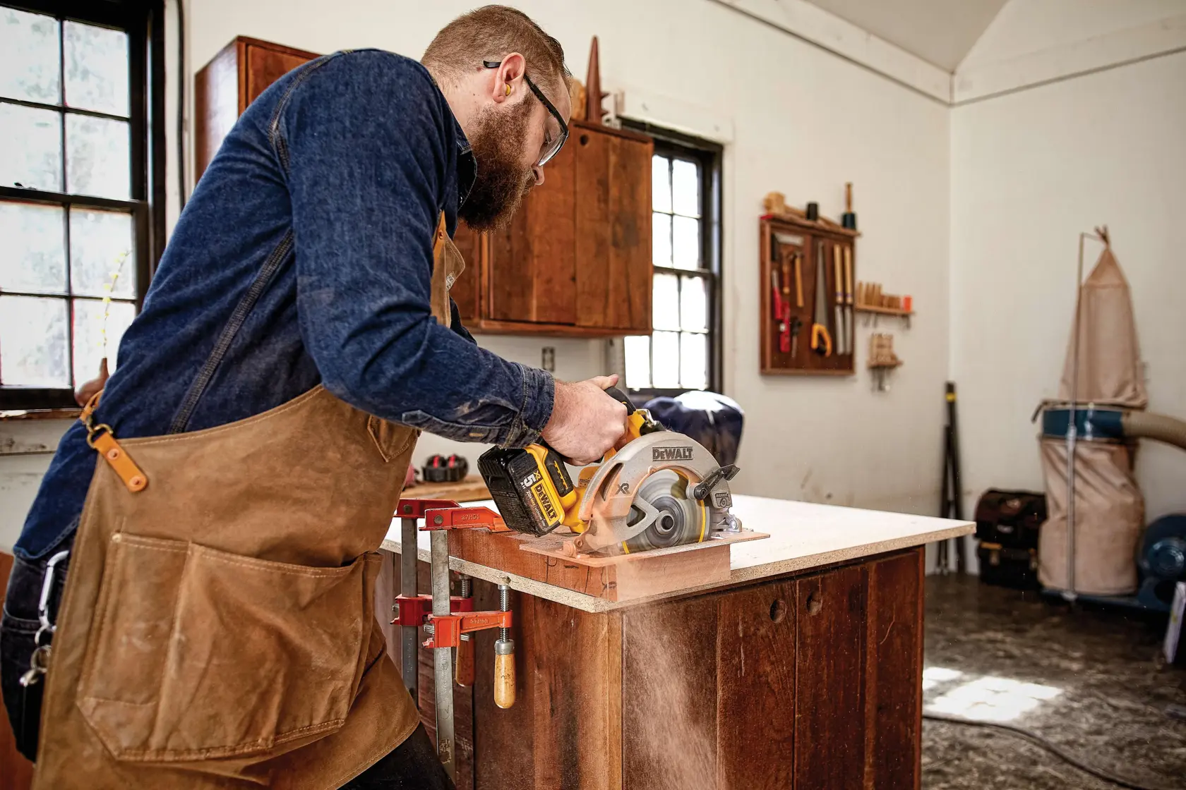 A person wearing a brown apron and blue shirt is using a DEWALT cordless circular saw (model DCS570B) to cut a piece of wood in a workshop with cabinets, workbench, and woodworking tools in the background.